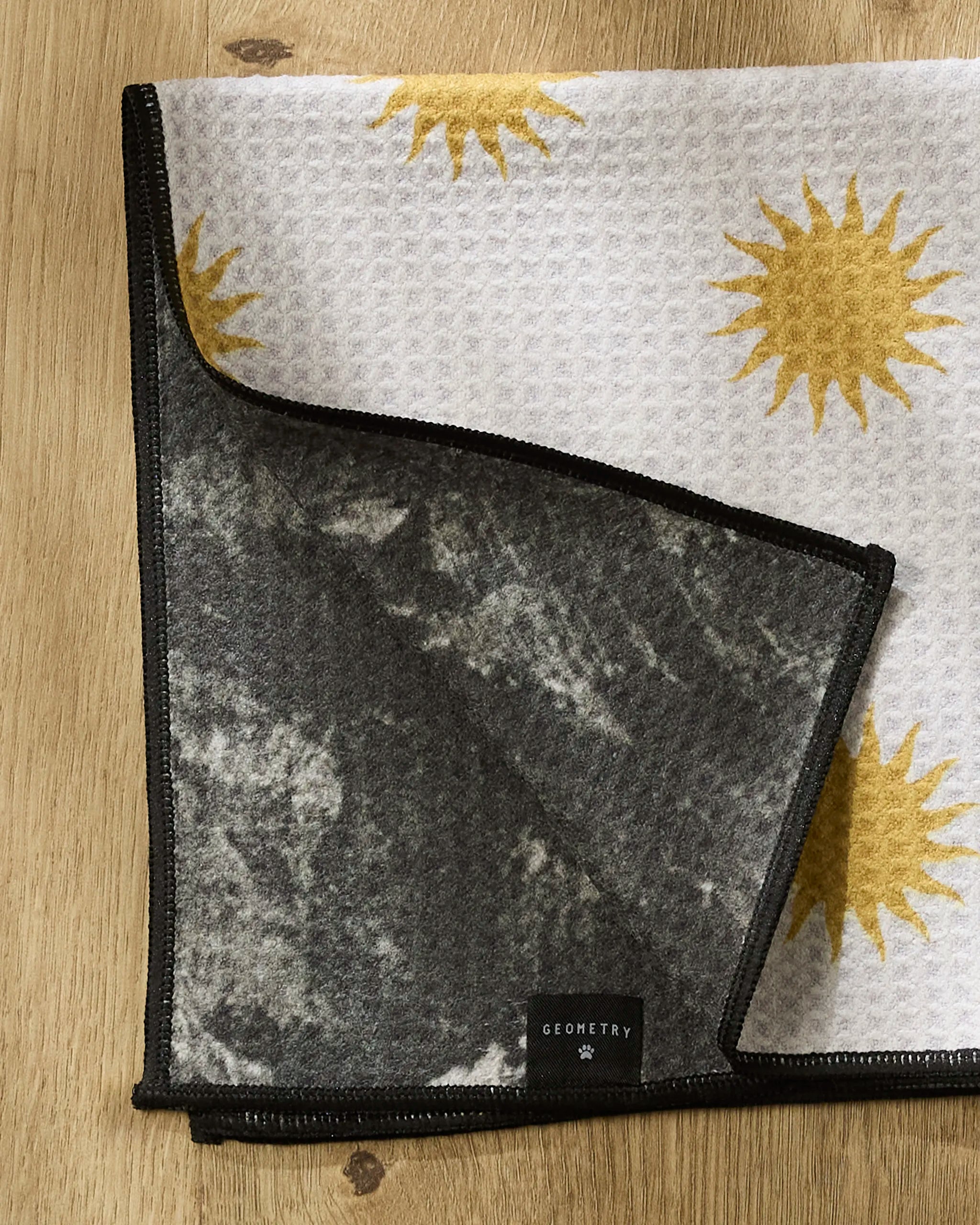 The Geometry Feeling Sunny dog towel features a white, waffle-textured surface with yellow sun prints, a dark gray marbled underside, and a black GEOMETRY tag, shown folded on a wooden surface.
