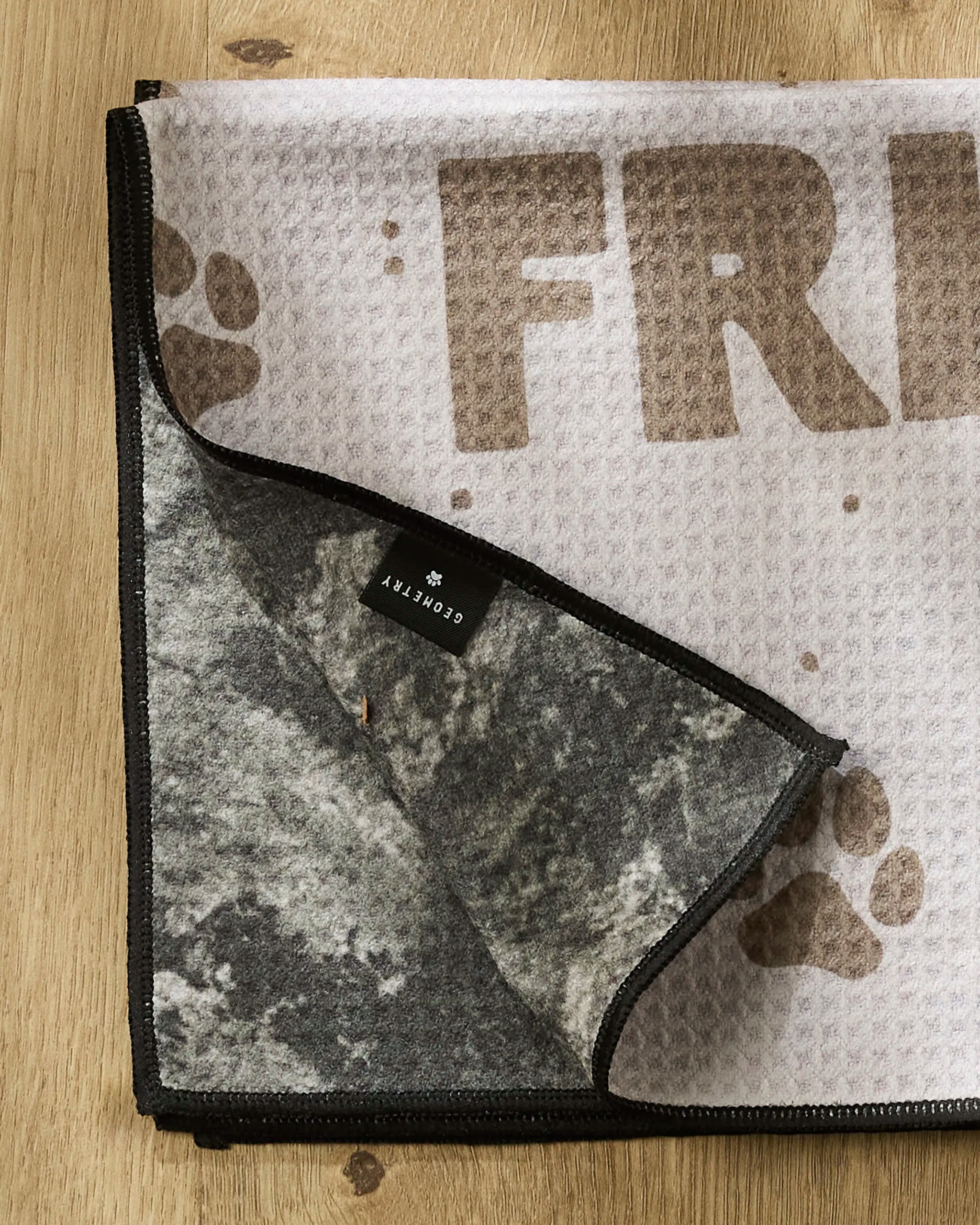 A folded Geometry Fur Ever Friends dog towel with a paw print design and "FRIEND" visible rests on wood. The flipped corner reveals a gray marbled underside and a black label.