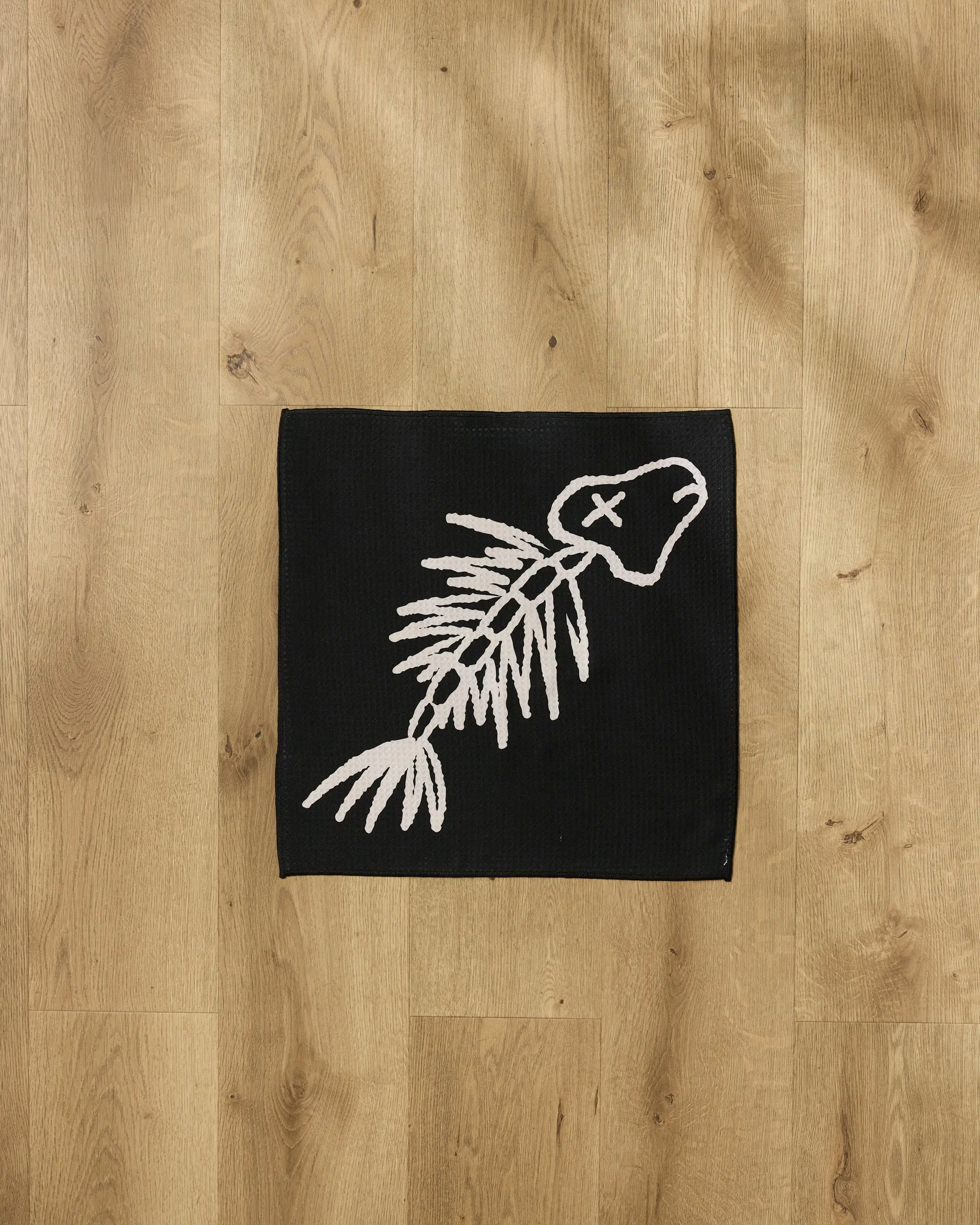 The Cole Strem "Gone Fishing" towel is a black recycled PET rectangle with a white fish skeleton (X for eye) design, shown on a wooden floor.