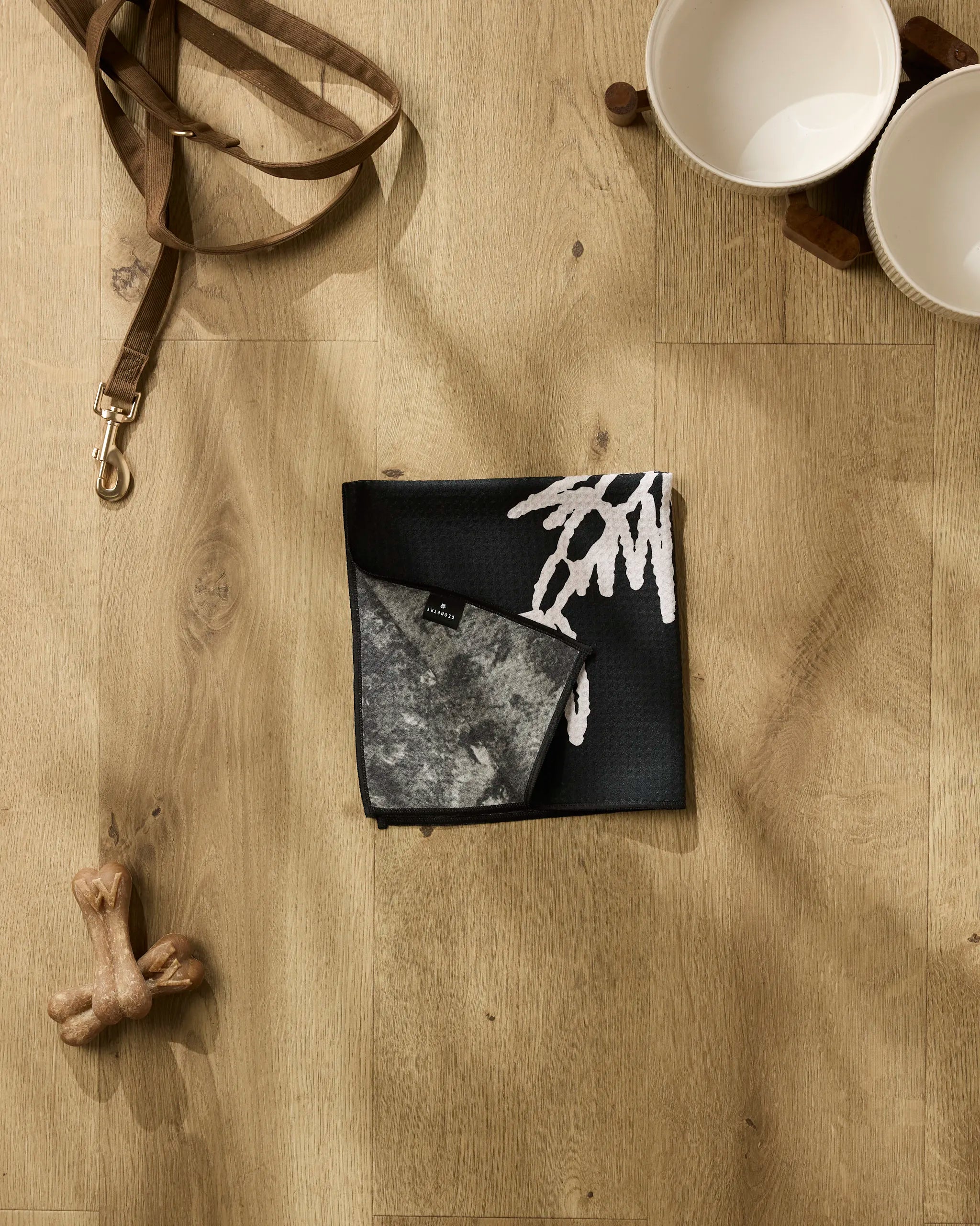 The Gone Fishing set by Cole Strem features a dog leash, two white bowls on a wooden stand, a folded black and gray pet bandana with a white design, two dog bones, and an ultra-absorbent towel on a light wood floor.