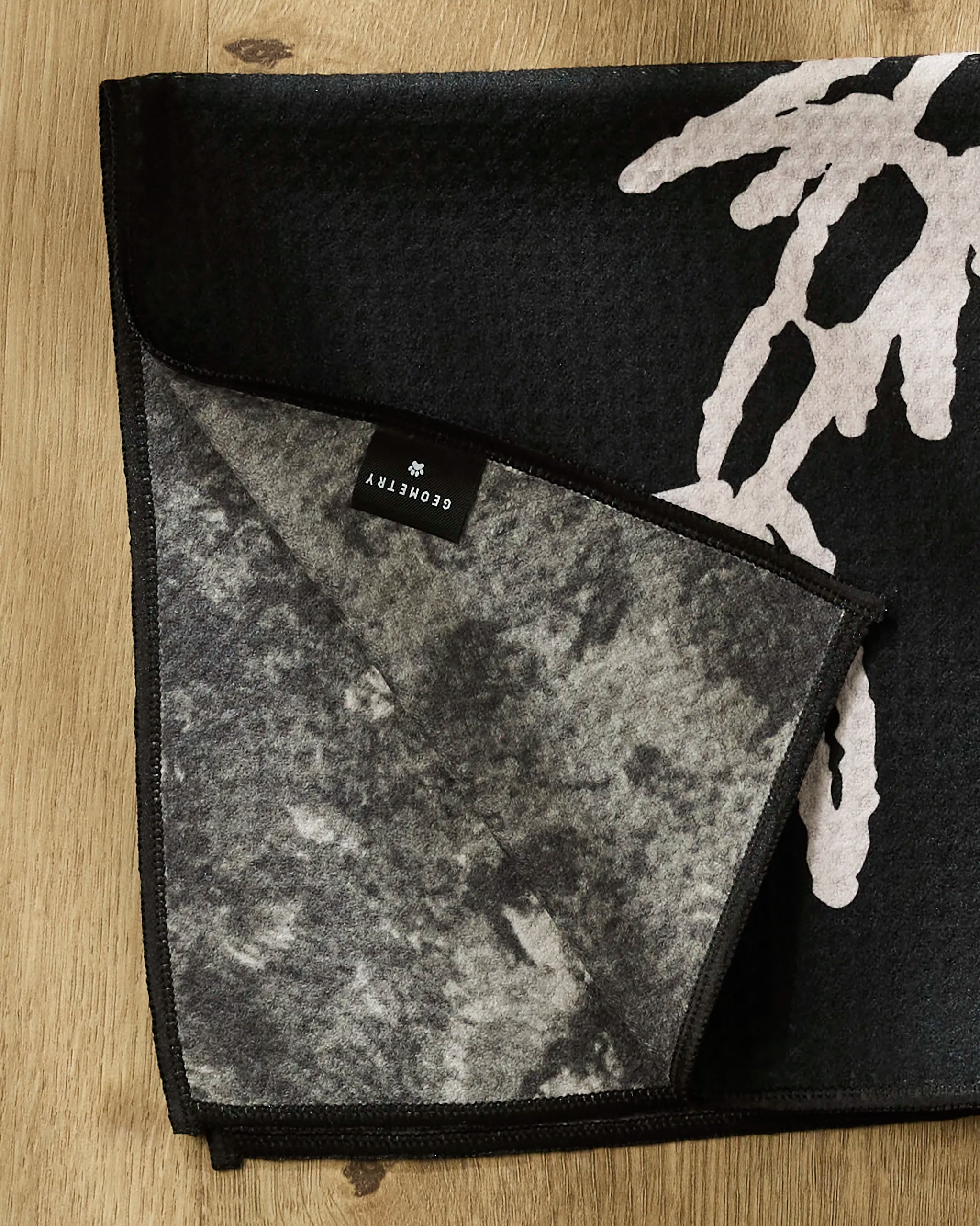 A close-up of the Gone Fishing ultra-absorbent dog towel by Cole Strem, in black and gray with a white abstract pattern, rests on wood. A folded corner reveals its textured underside and a tag labeled Cole Strem.