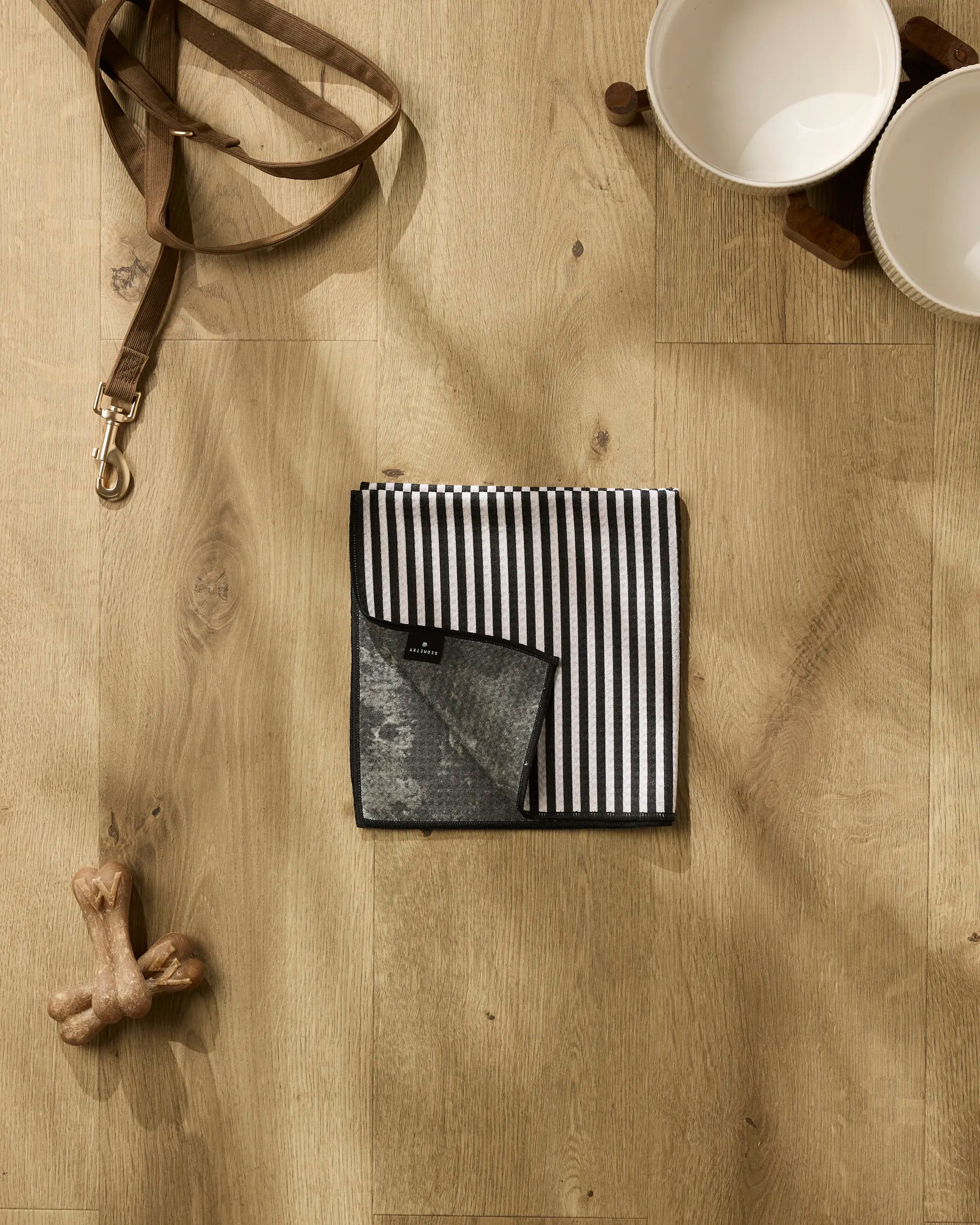 A Geometry Good Pup recycled PET towel with black and white stripes is folded on a wooden floor beside two ceramic pet bowls, a brown leather leash, and a bone-shaped dog toy.