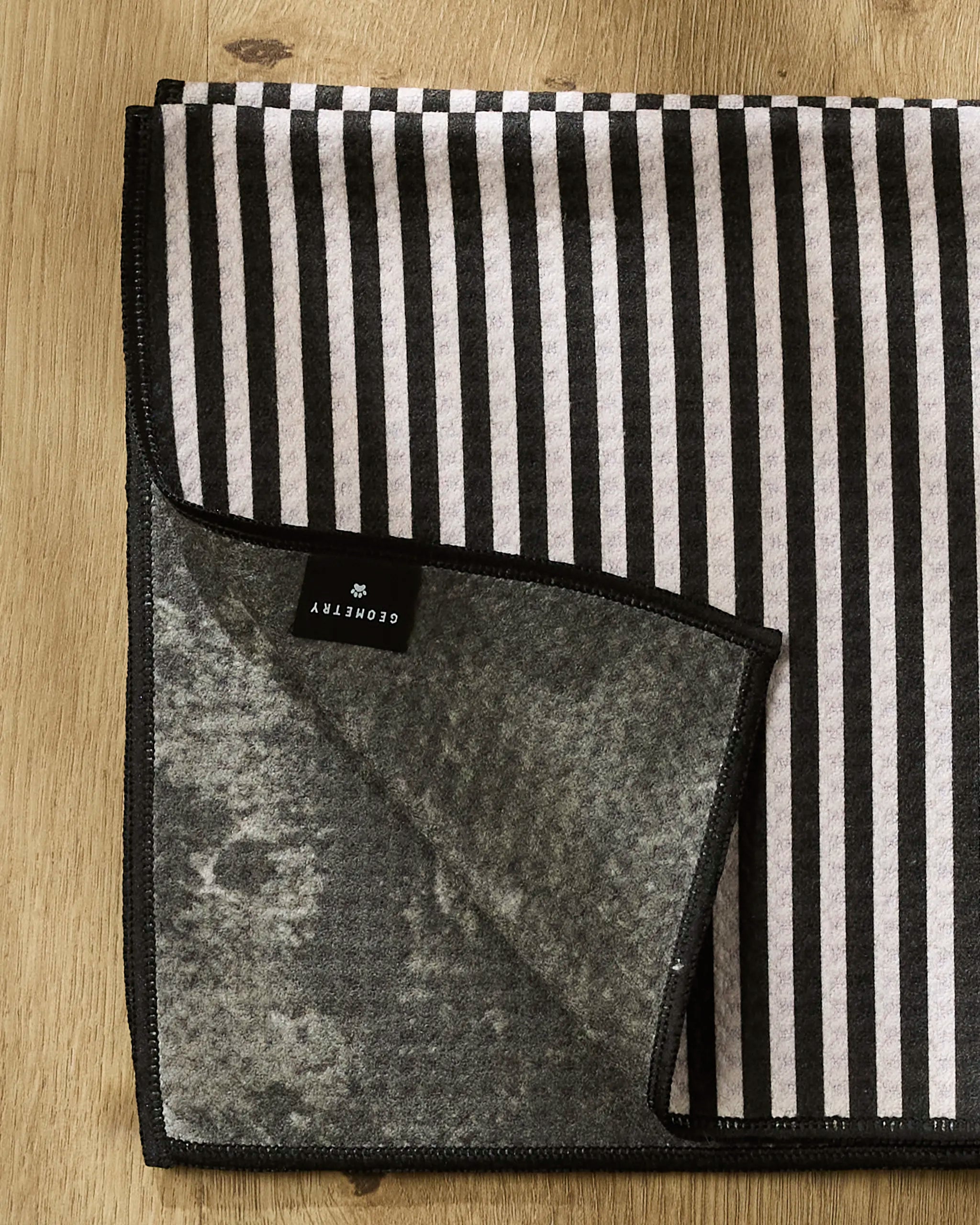 A folded black and white striped Good Pup towel by Geometry with black edging rests on wood; a flipped corner reveals its textured gray underside and a black "Geometry" label.