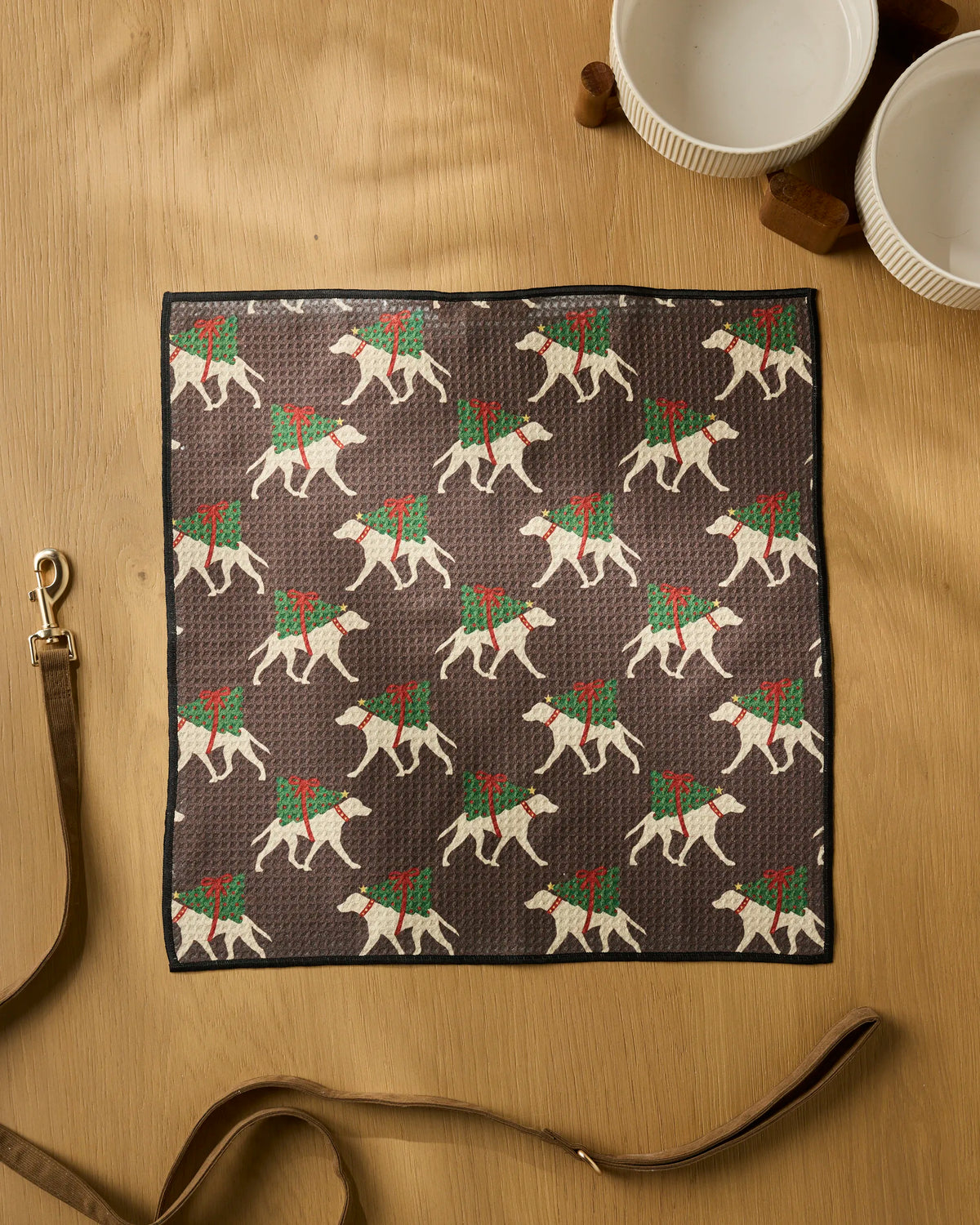 The Geometry Holiday Helpers dog towel, an ultra-absorbent brown square cloth with a festive pattern of white dogs in green and red scarves, is displayed on a wooden surface alongside a dog leash, two white bowls, and brown bone-shaped dog treats.