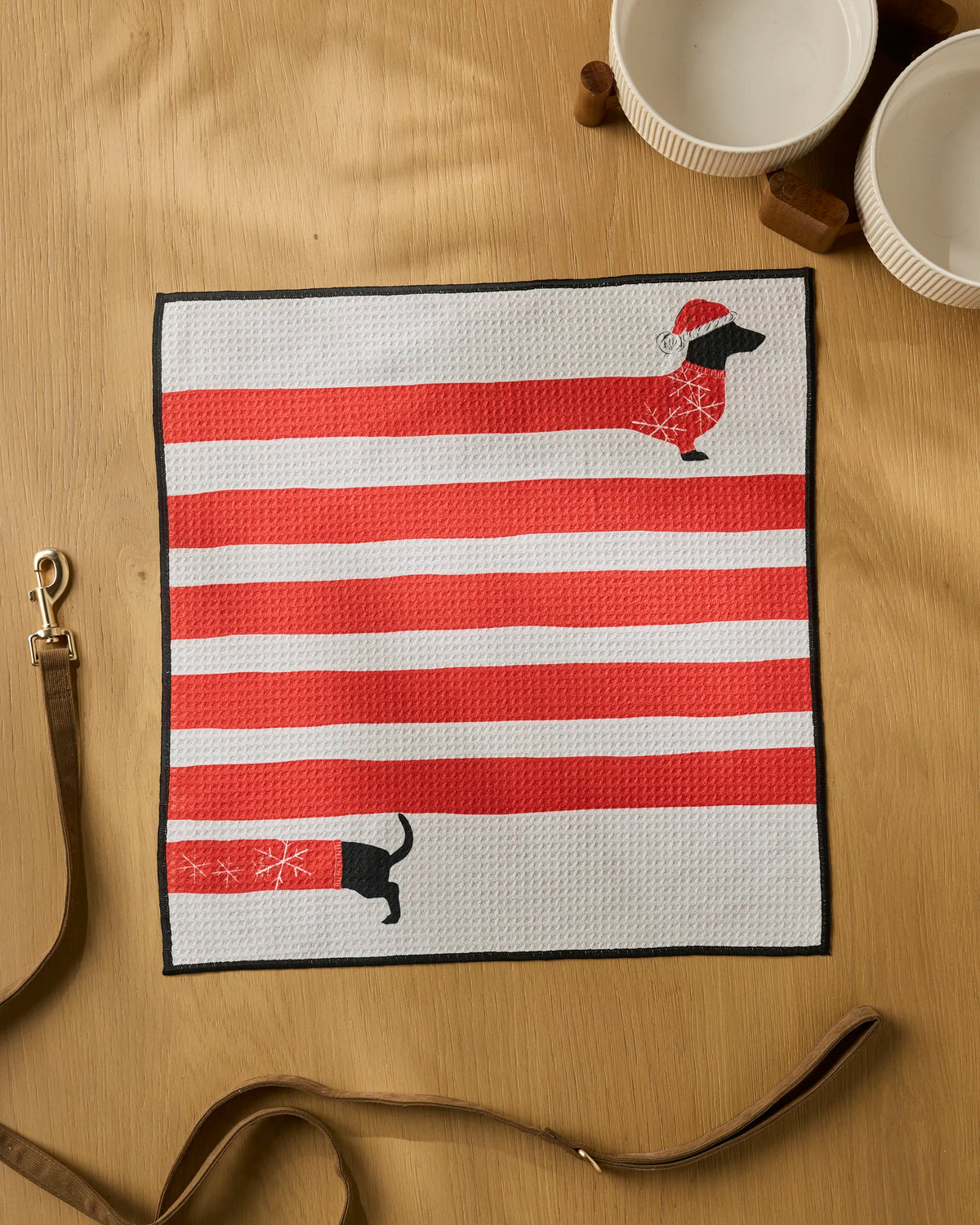 The Geometry Holiday Stretch mat, featuring red and white stripes with dachshund dogs—one sporting a Santa hat and scarf—serves as an ultra-absorbent pet towel on a wooden table alongside two white bowls, a brown dog leash, and nearby wooden accents.