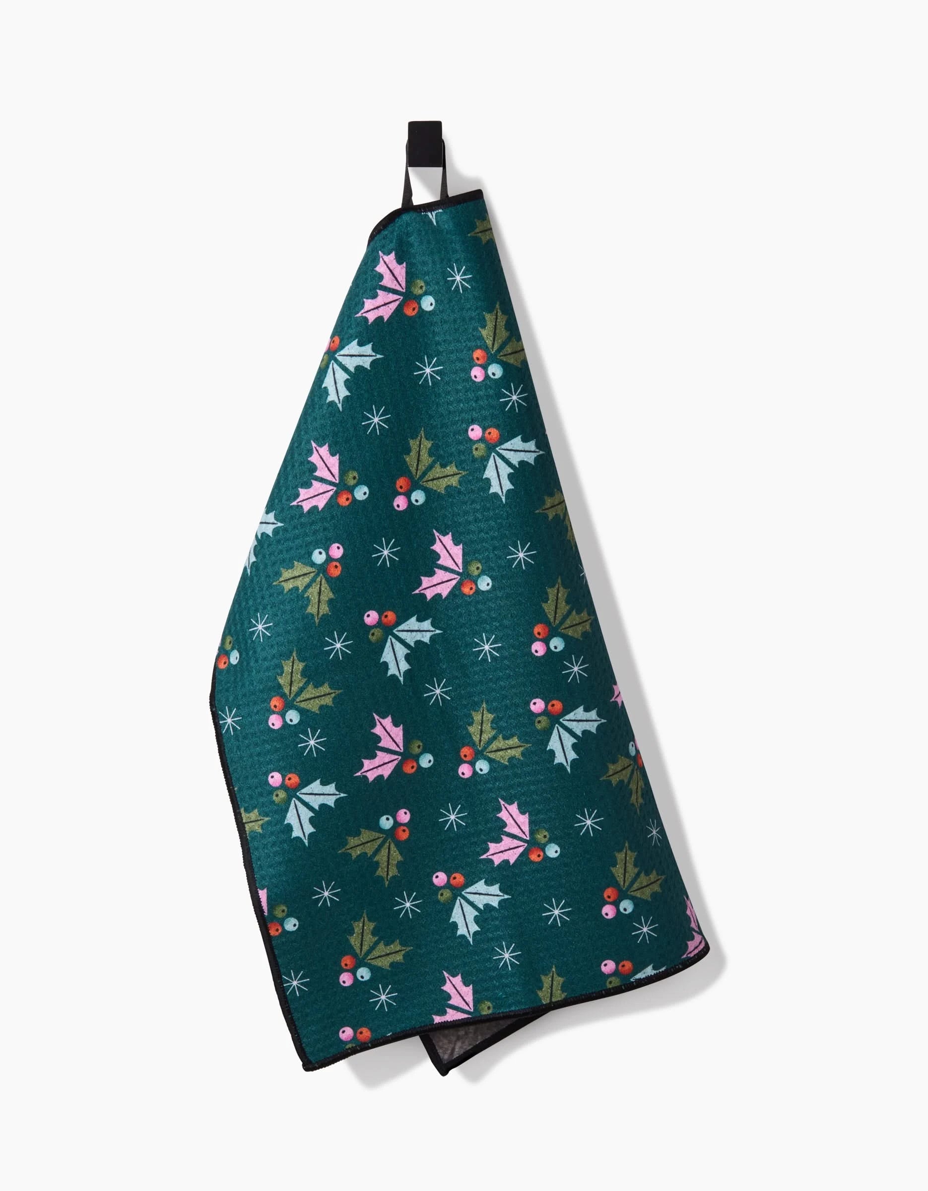 The Megan Roy Holly Berries Paw Towel, in dark green with pink and green holly leaves, red berries, and white snowflakes, hangs from a black loop against a white background.