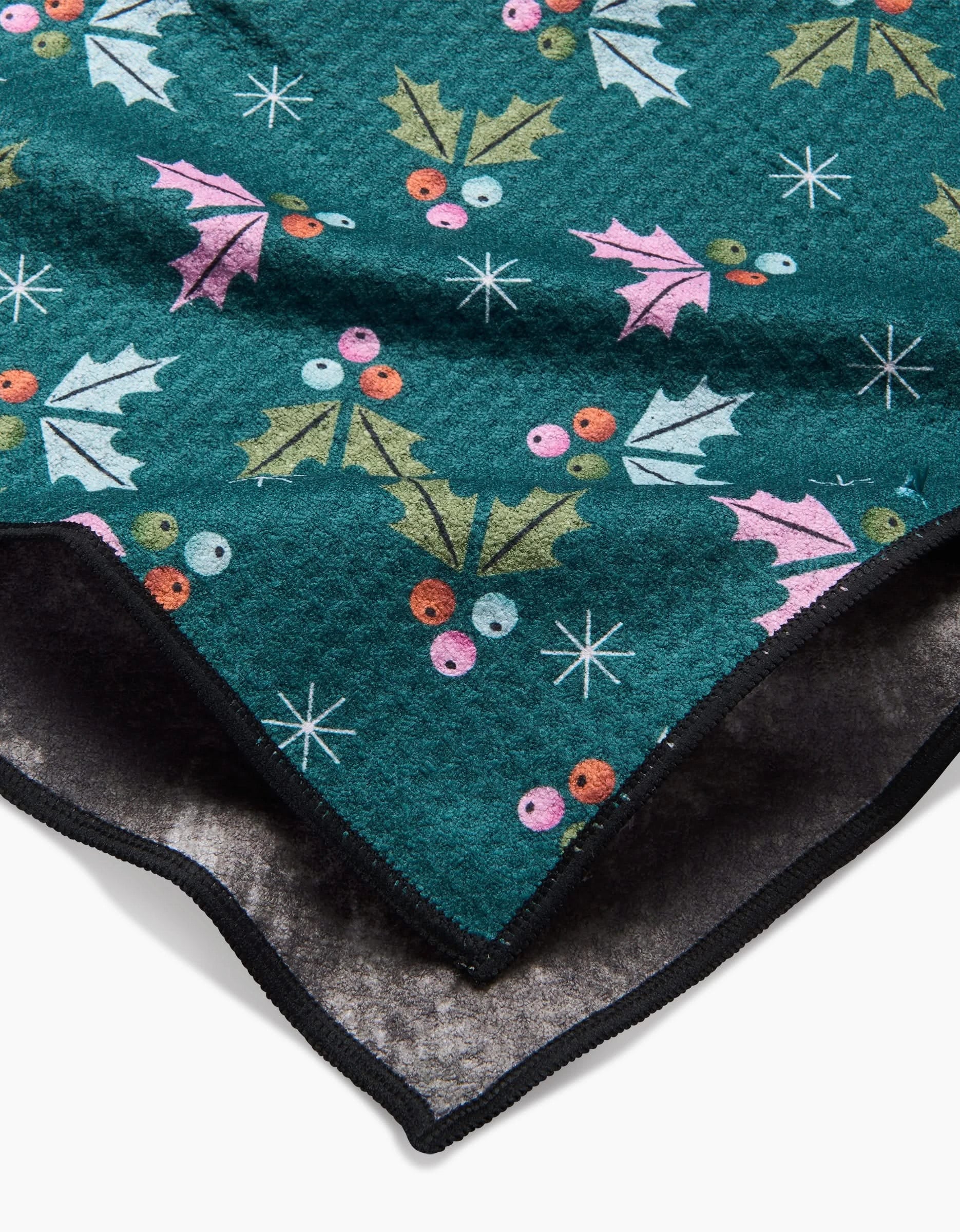 A close-up of Megan Roys Holly Berries Paw Towel, a soft green fleece with festive holly leaves, red and pink berries, white stars, ultra-absorbent dark underside, and black-trimmed edges.