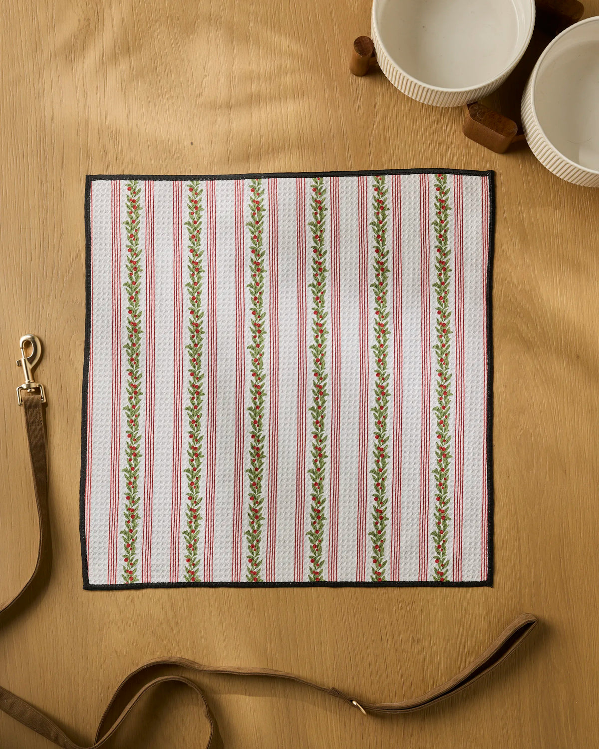 A square cloth from Geometry, called Holly Vine, featuring red and green vertical floral stripes, lies on a wooden table. Two white bowls and a brown leather leash are nearby, highlighting this ultra-absorbent dog towel’s stylish design and practicality for your pet’s needs.