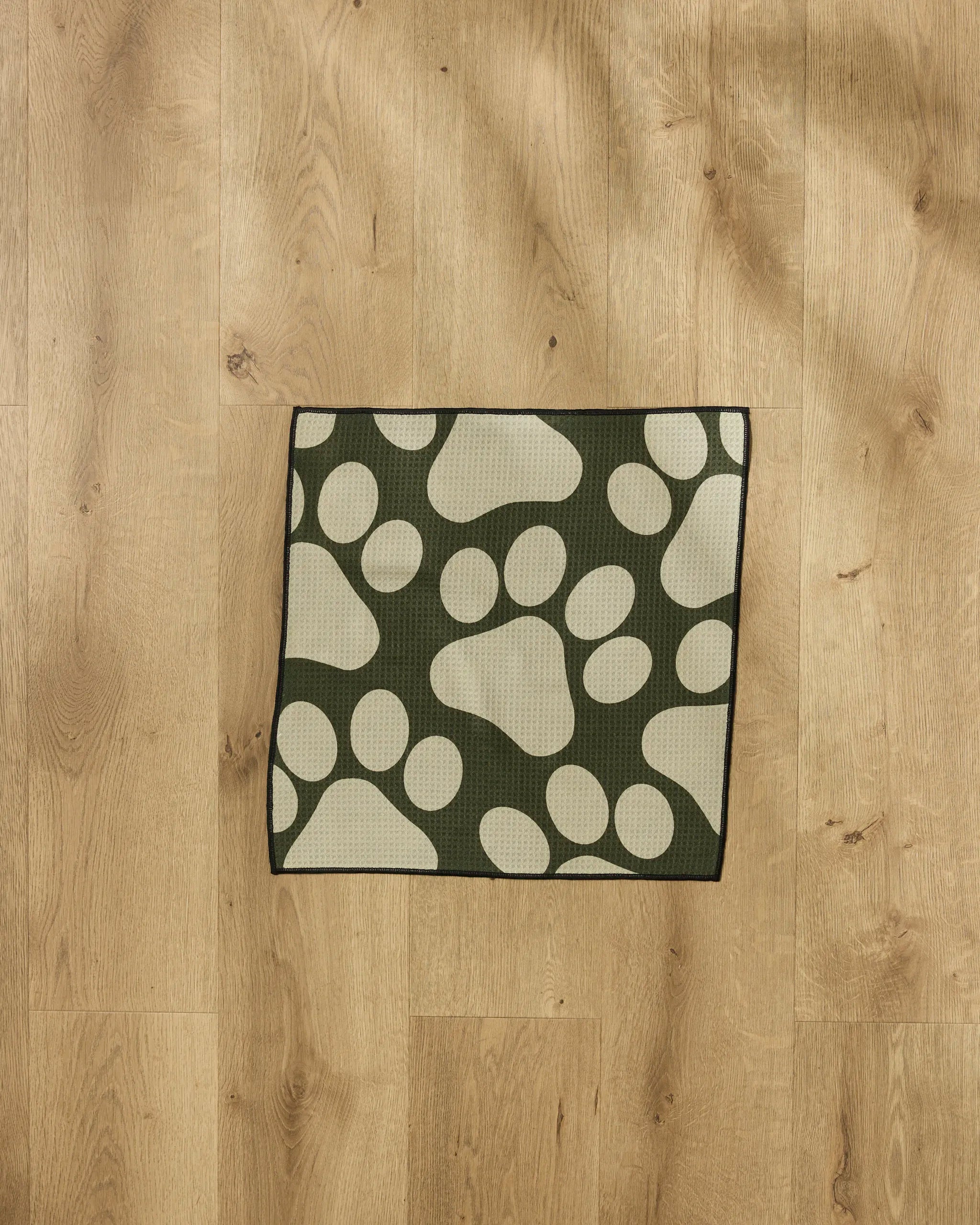 The Geometry Paw Prints mat, featuring large cream paw prints on green, sits on a light wood floor—perfect for catching messes after using an ultra-absorbent dog towel.