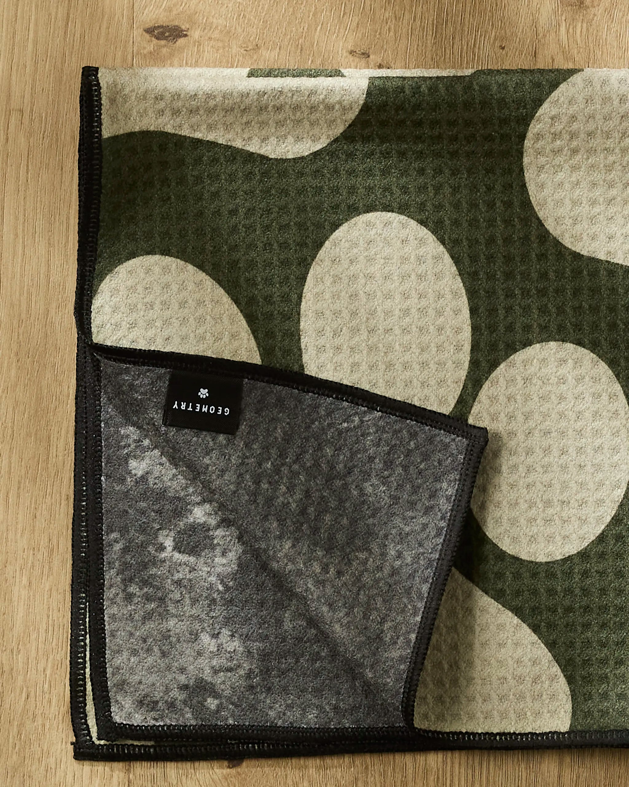 The Geometry Paw Prints towel features large cream circles on olive green with black trim, shown folded on wood. A corner flips to reveal the soft gray underside and sewn black label.