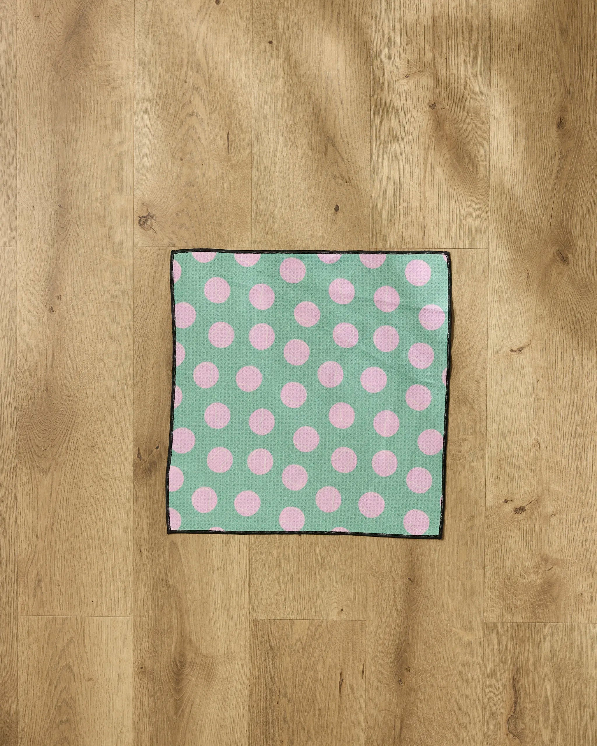 A "Pink Ball" recycled pet towel by Geometry, featuring large pink polka dots and a black border, is laid flat on a light wooden floor.