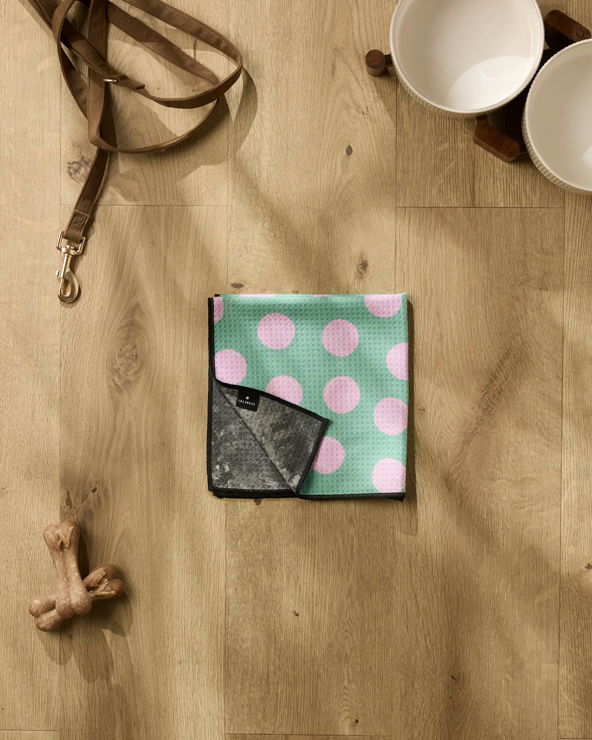 A Pink Ball by Geometry sits on a wooden floor next to a brown dog leash, two white bowls on a wooden stand, and a dog bone toy.