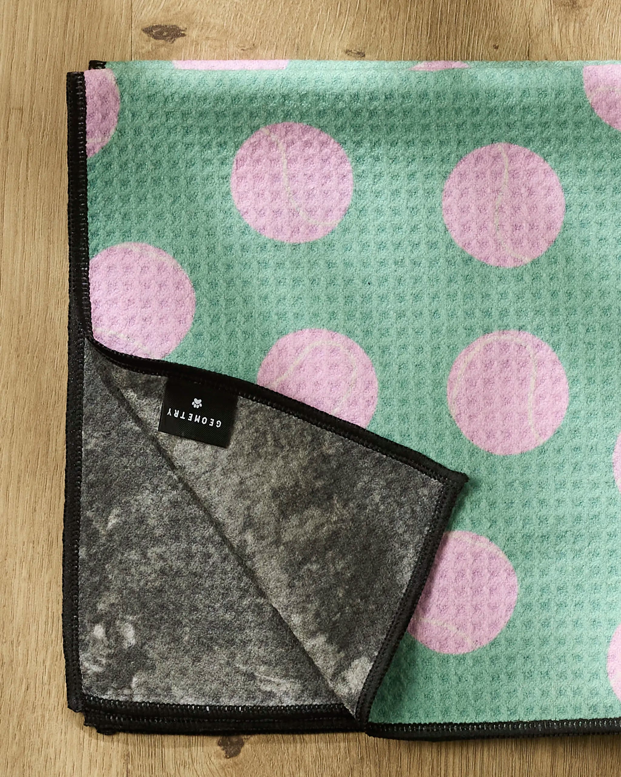 The Geometry Pink Ball towel, made from recycled PET, features a green surface with large pink polka dots, black trim, a gray underside, and a black label on the corner. This ultra-absorbent dog towel is shown folded on a wooden surface.