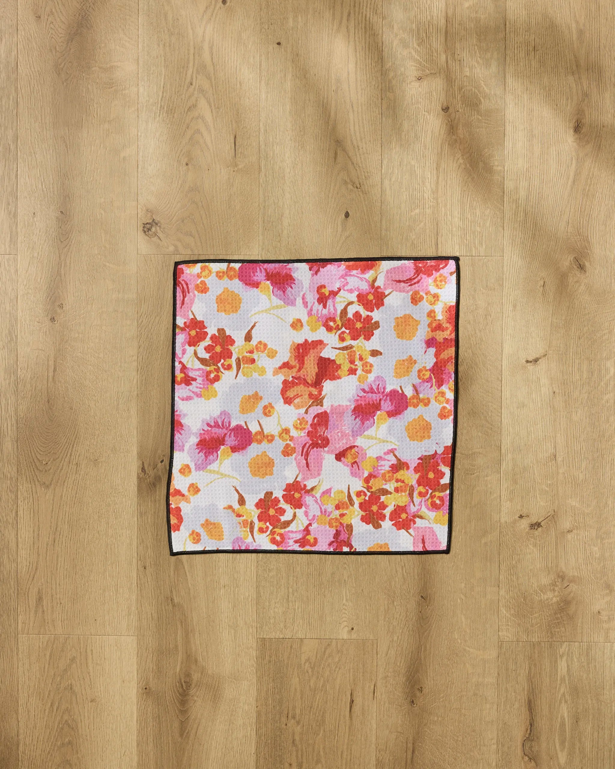 The Geometry Pup Florals Paw Towel, featuring a vibrant red, orange, yellow, and pink floral design with a dark border, lies on a light wood floor. This ultra-absorbent towel combines charm and function for pet care.