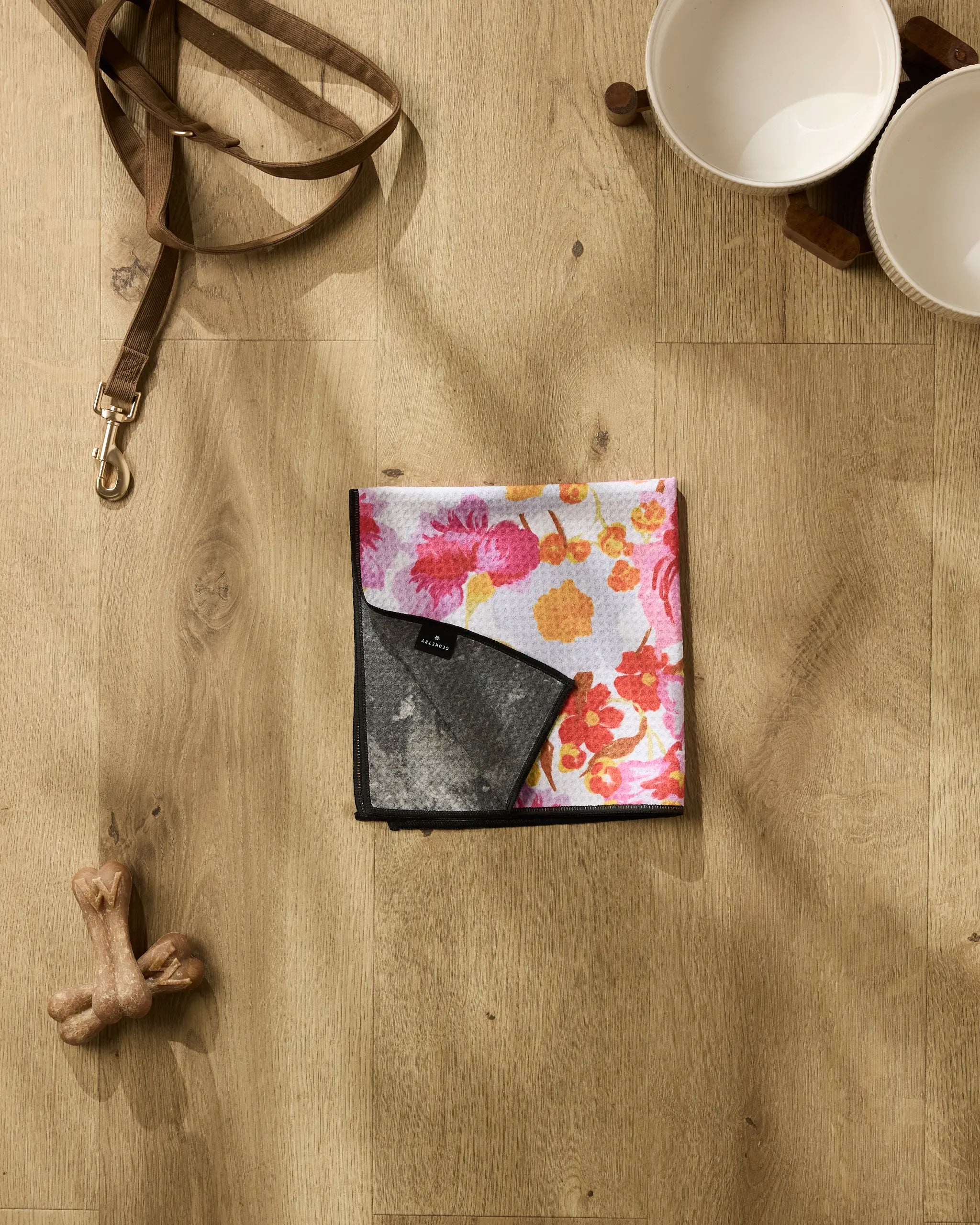 The Geometry Pup Florals dog bandana is displayed on a wooden floor beside a brown leash, two white pet bowls on a stand, an ultra-absorbent dog towel, and a bone-shaped chew toy.