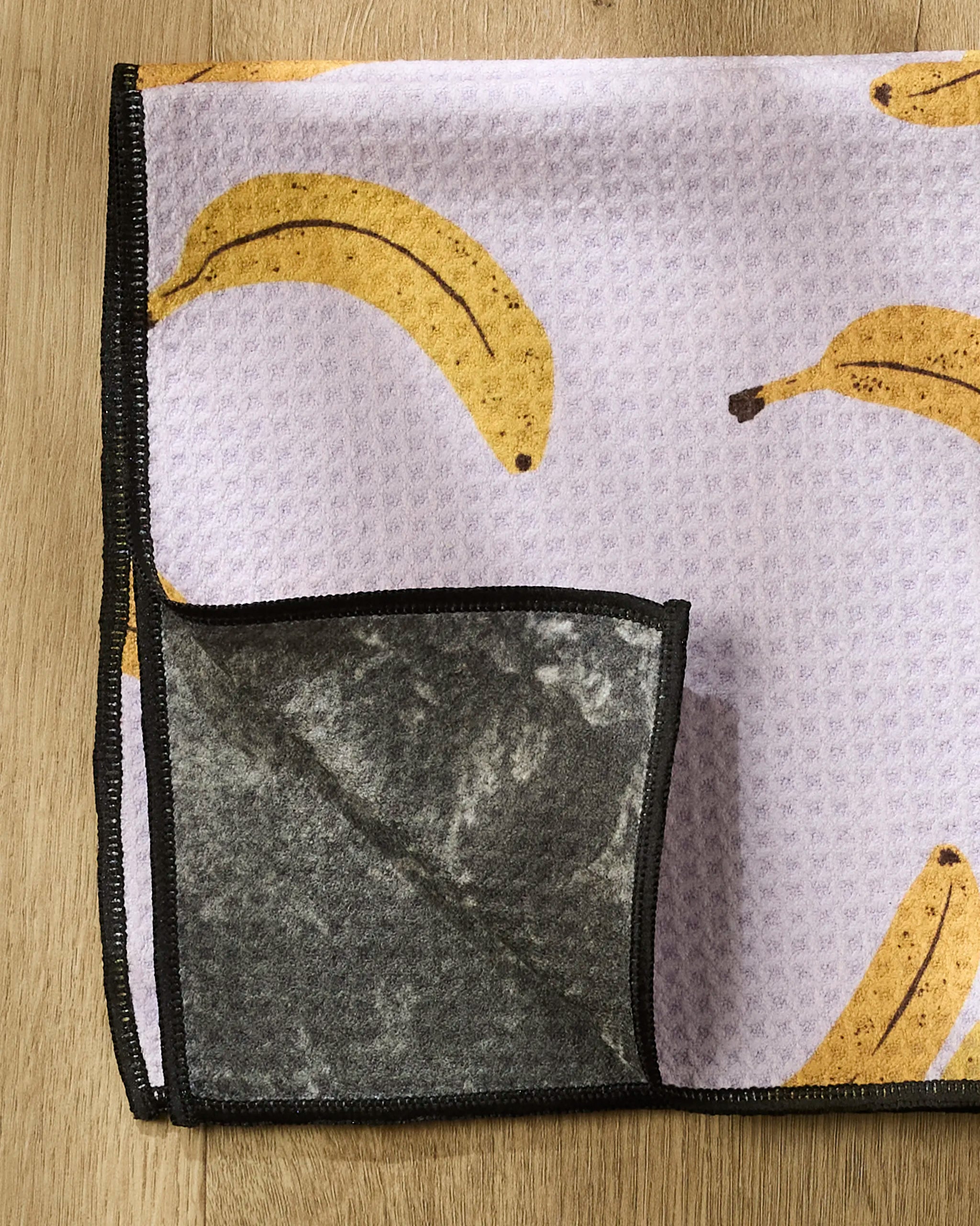 The Geometry Pupnana towel, made from recycled PET, features a light purple banana print front and a dark gray textured underside. It's shown on a wooden surface with one corner folded to display both sides.