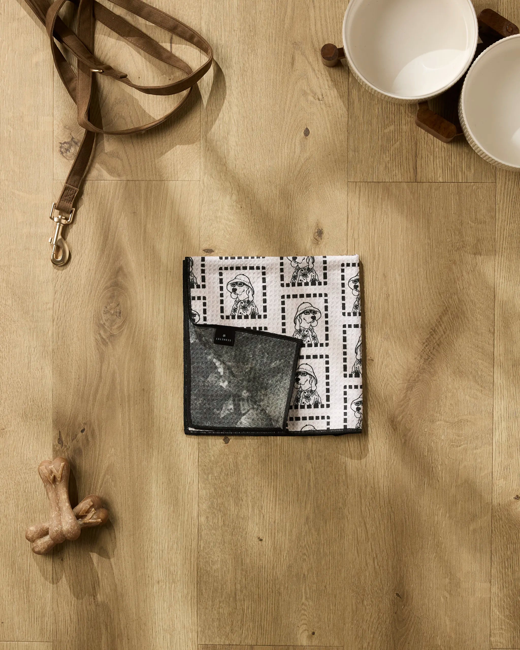 A flat lay features Geometry's Ready For Summer Doggy set: a leash, patterned bandana, ultra-absorbent dog towel, bone-shaped toy, and two white bowls on a wooden stand.