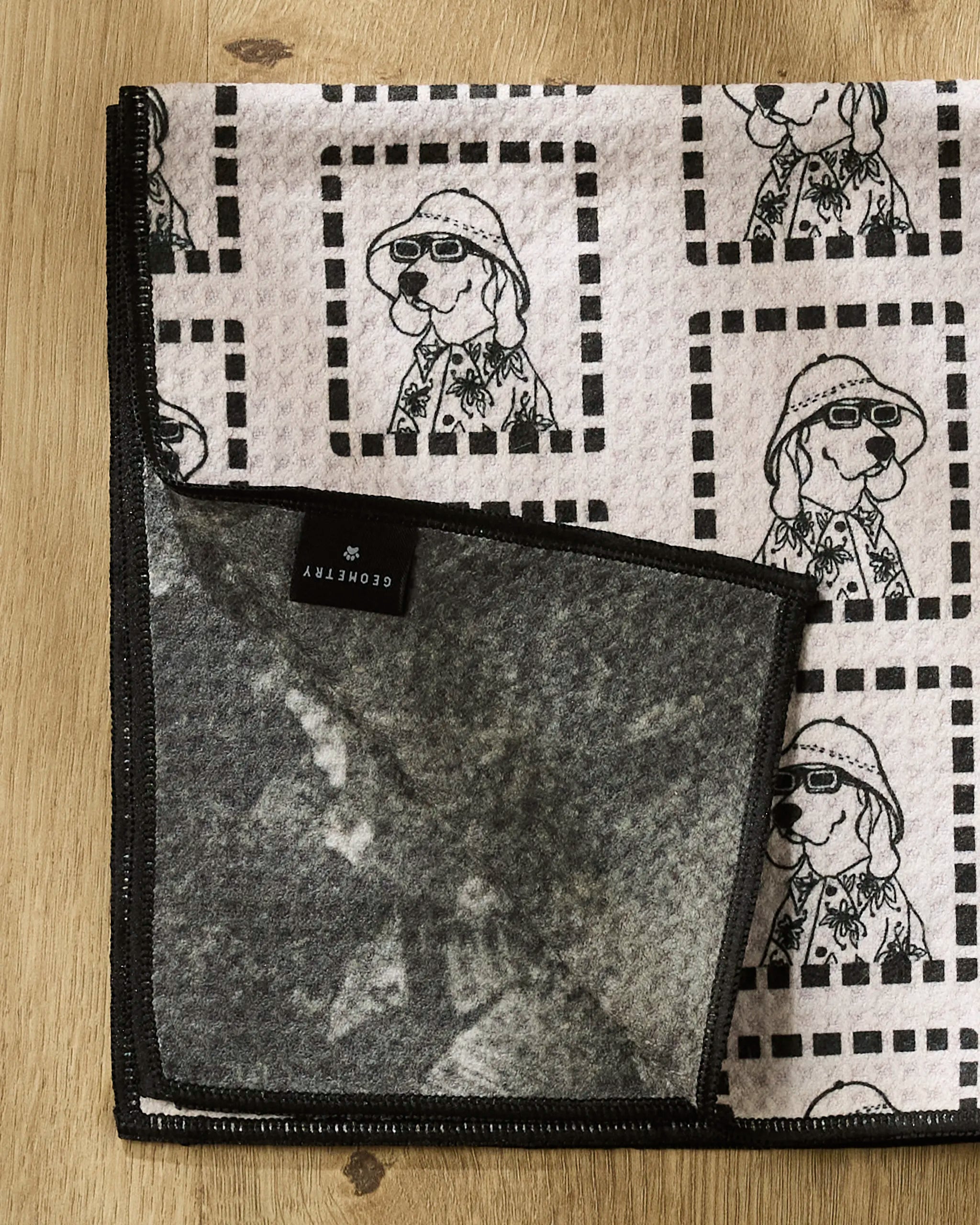 The Geometry "Ready For Summer Doggy" Paw Towel, featuring cartoon dogs in hats and glasses with square borders, is folded on a wooden surface. A black-edged corner shows the gray, fuzzy underside and a small black label.
