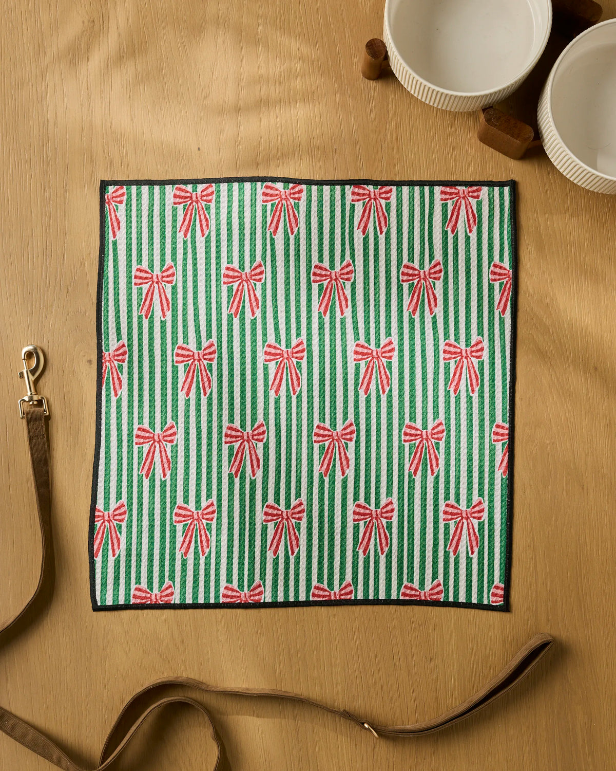 The Ribbon Toss dog towel by Geometry, featuring green and white stripes with red bows, is laid flat on a wooden surface alongside a leather strap, two white bowls, and a small wooden item.