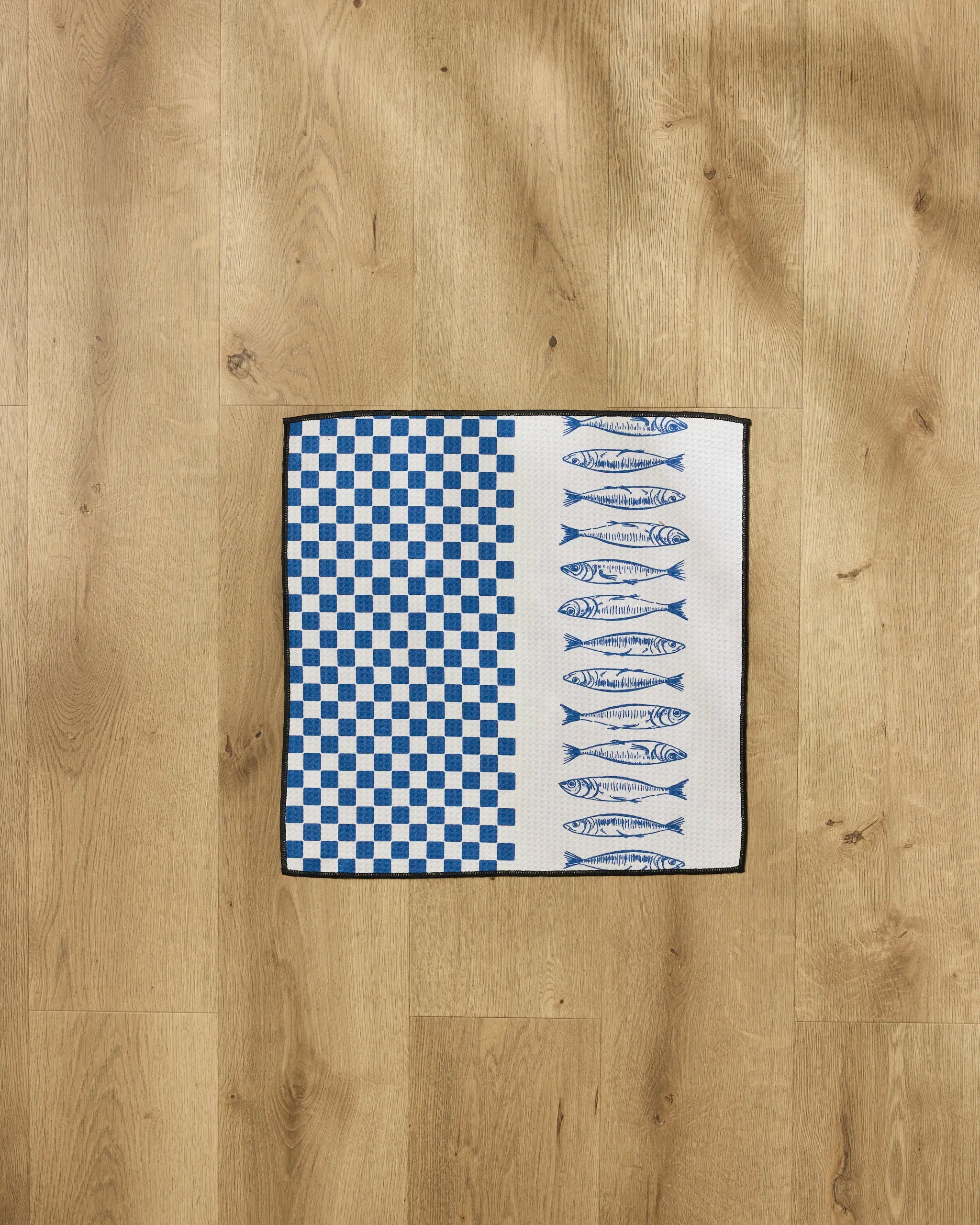 The Sardine Check Blue by Hufton Studio is a white recycled PET towel featuring a blue checkerboard pattern on the left and rows of blue fish on the right, displayed on a light wooden floor.