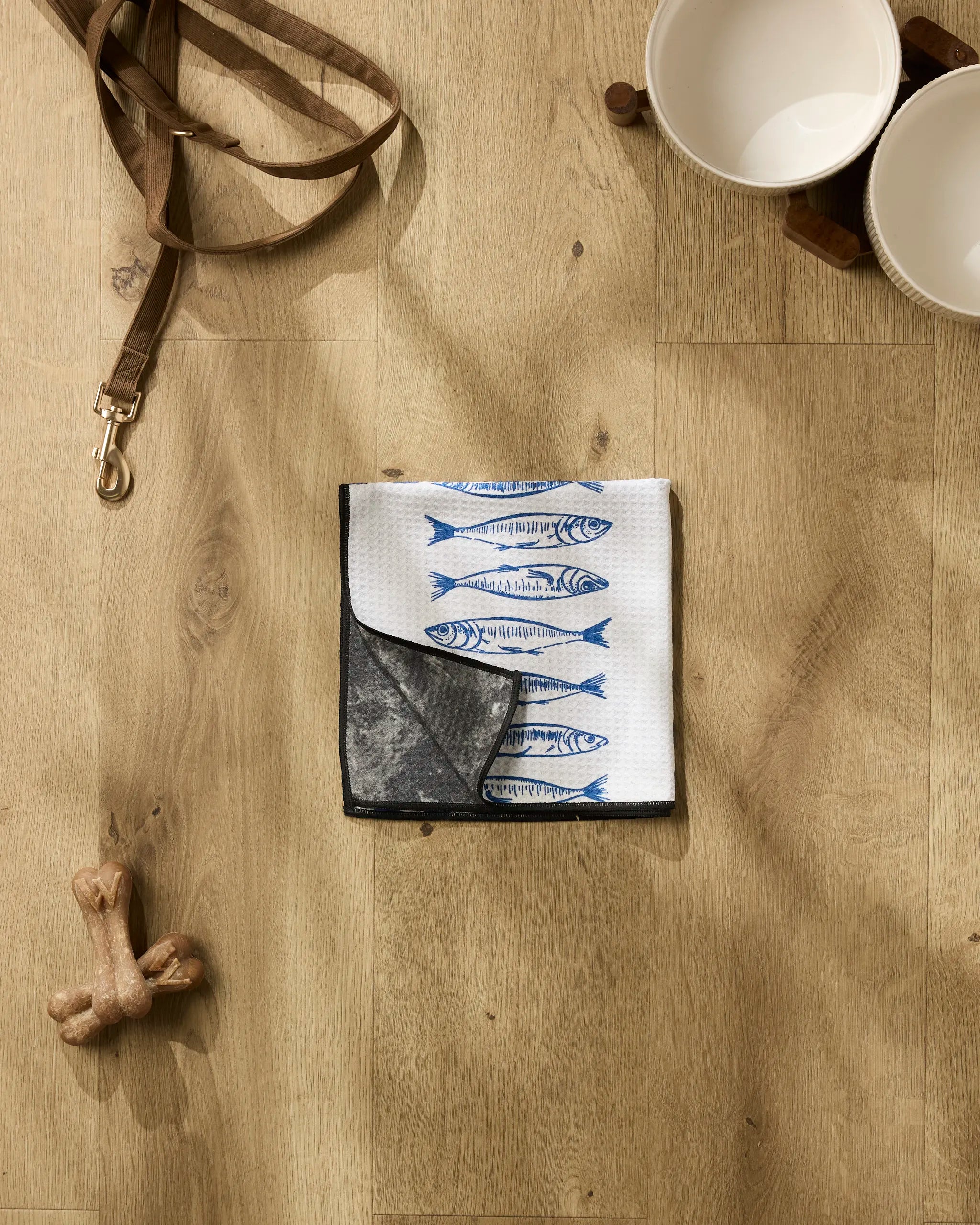 The Sardine Check Blue ultra-absorbent dog towel by Hufton Studio, a dog leash, two white pet bowls, and a bone-shaped toy are neatly arranged on a light wooden floor.