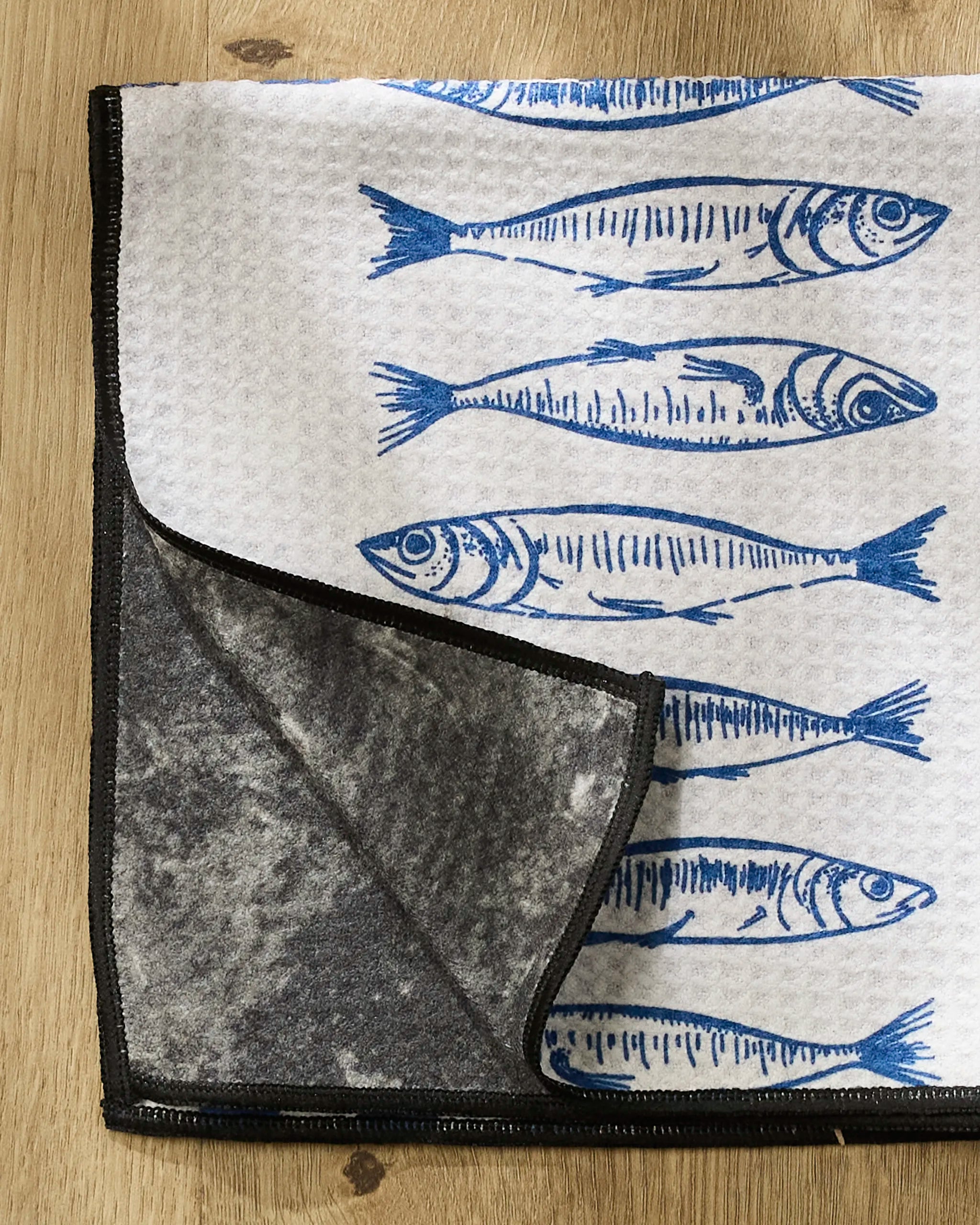 The Sardine Check Blue by Hufton Studio is a folded white kitchen towel with blue fish illustrations and black trim, shown on light wood. Its flipped corner reveals a textured dark gray underside—perfect as an ultra-absorbent paw towel for pets.