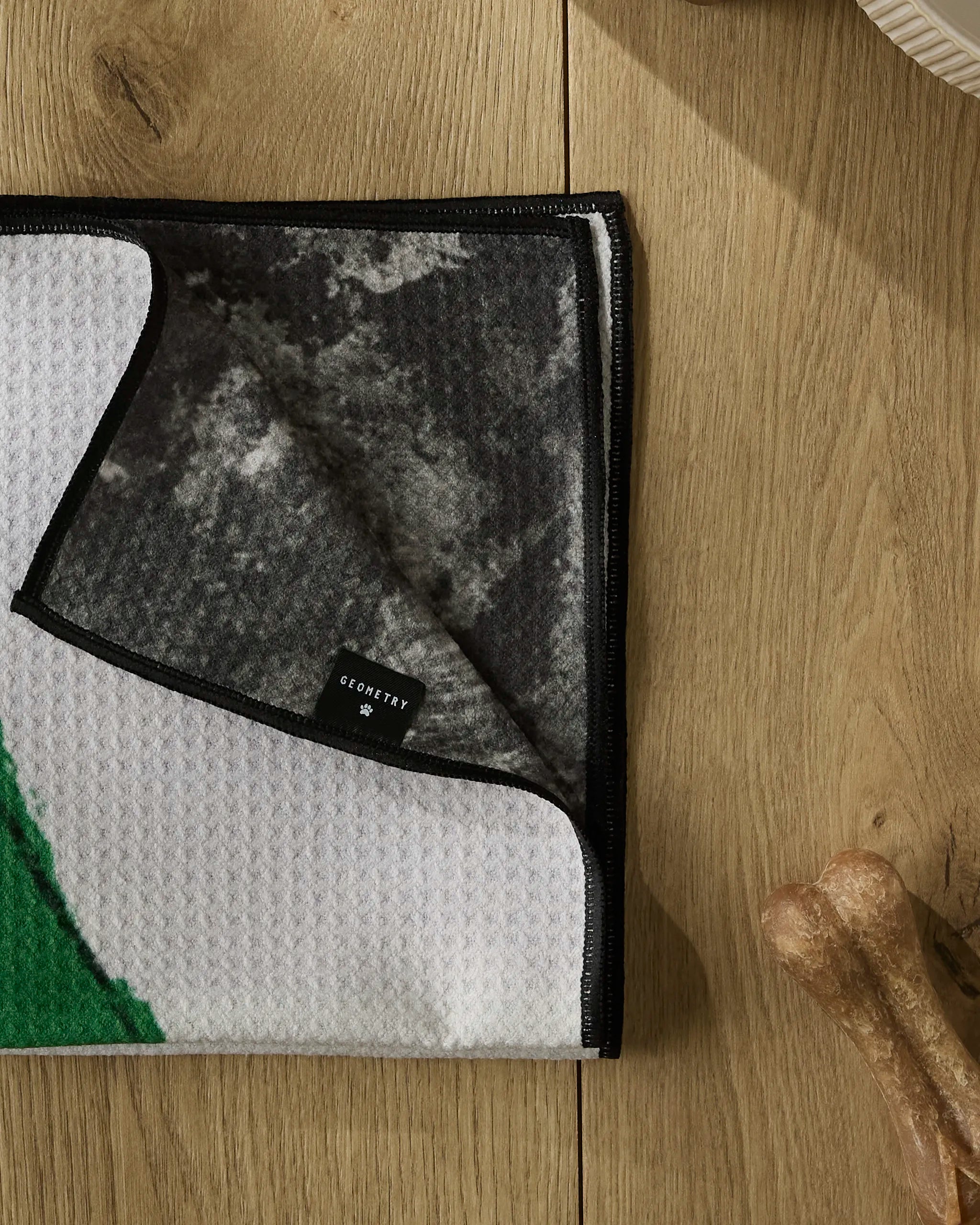 A close-up of the Silly Stocking paw towel by Geometry, featuring a green and white geometric design and a small black GEOMETRY tag, lies on a wooden surface with a bone-shaped dog treat in the corner.