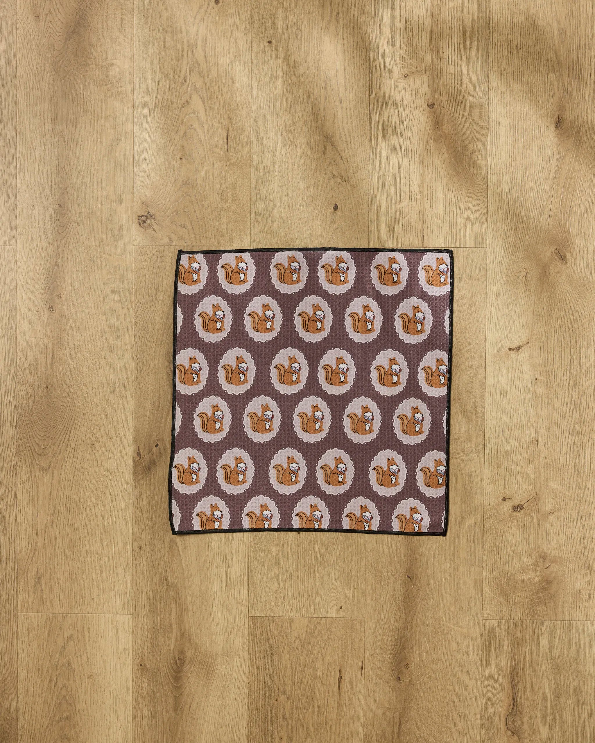 The Megan Roy "Squirrels Brown" mat features a playful squirrel pattern on a brown background and rests on a light wooden floor—perfect as an ultra-absorbent dog or paw towel for your furry friend.