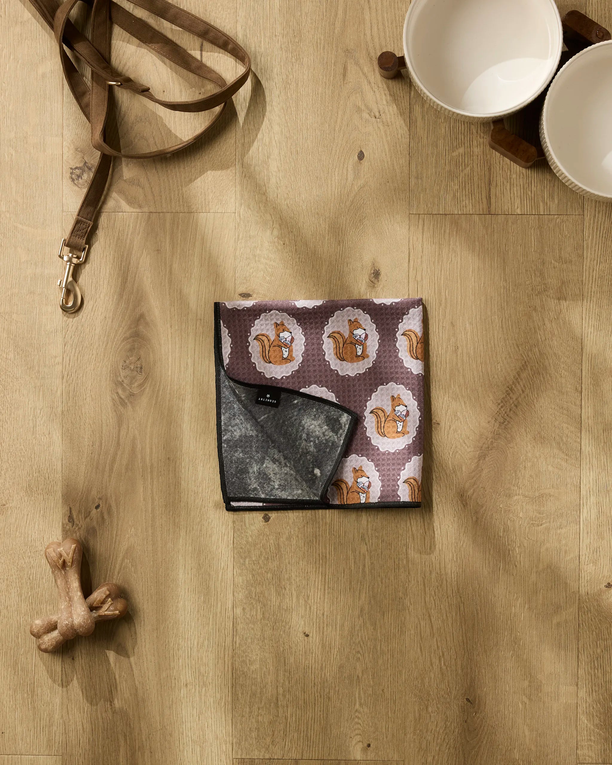 The Megan Roy Squirrels Brown dog leash, two white bowls on a wooden stand, a dog bone, and an ultra-absorbent towel with a cartoon squirrel print are displayed on a light wood floor.