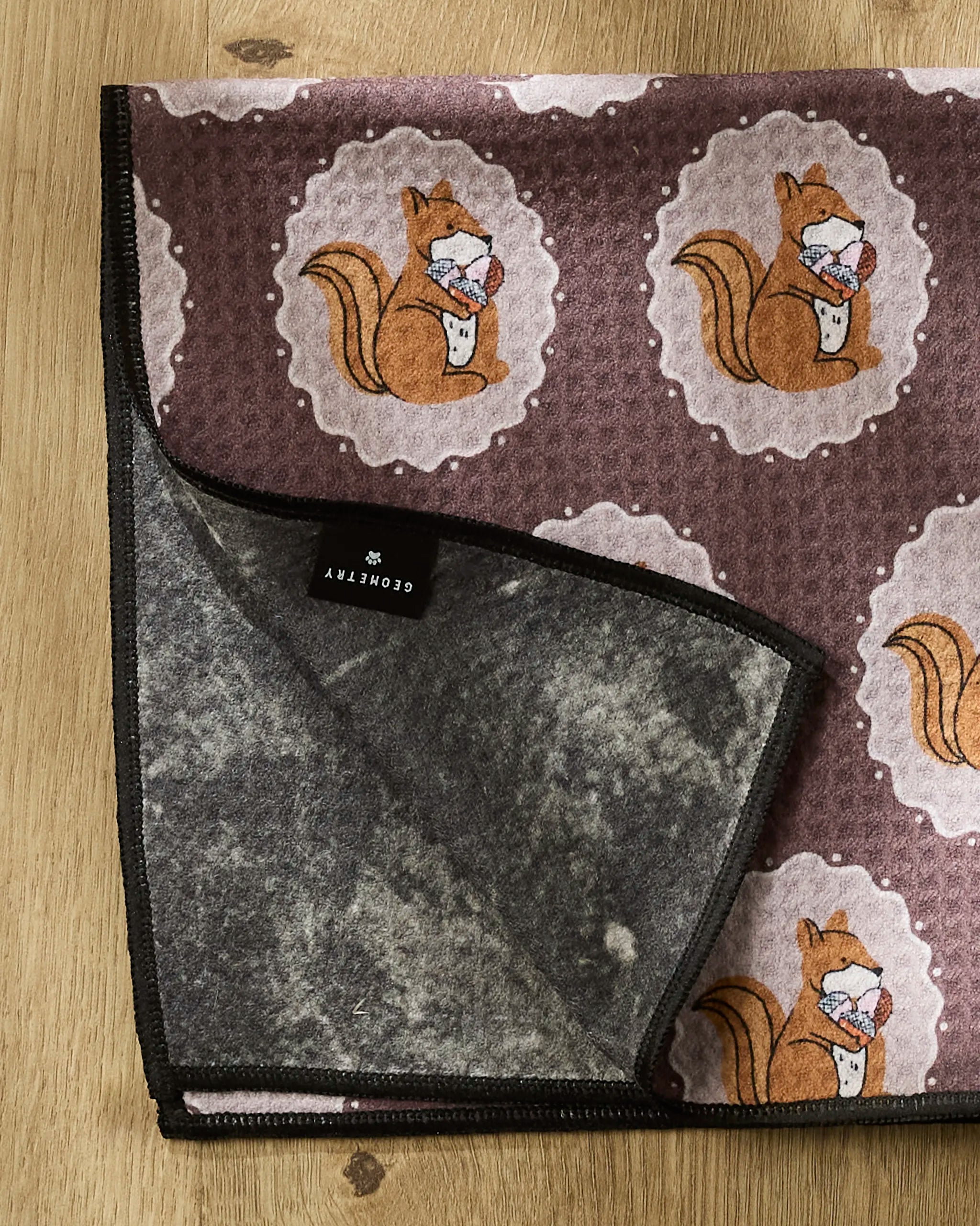 A brown and gray recycled PET towel called "Squirrels Brown" by Megan Roy, featuring a squirrel pattern, is shown on wood with one corner flipped to reveal the underside and a black GEORGETRY label.
