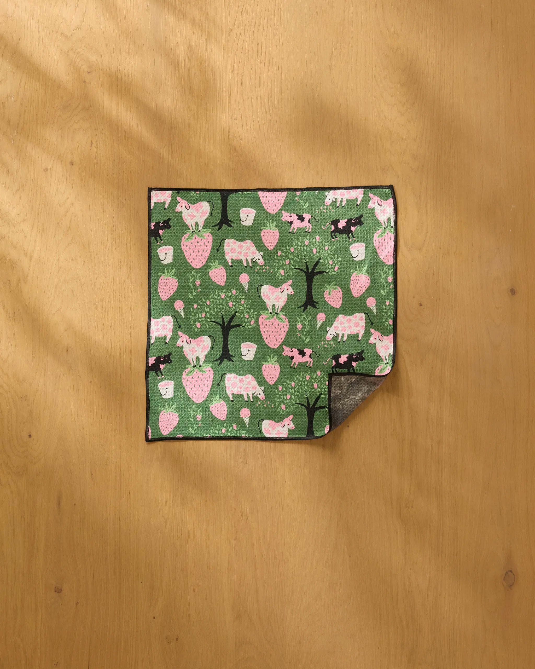 A green rectangular ultra-absorbent dog towel, "Strawberry Cows" by Louise Pretzel, features pink and white animal, tree, and mushroom patterns. It lies on a light wooden surface with one corner folded to show the underside.