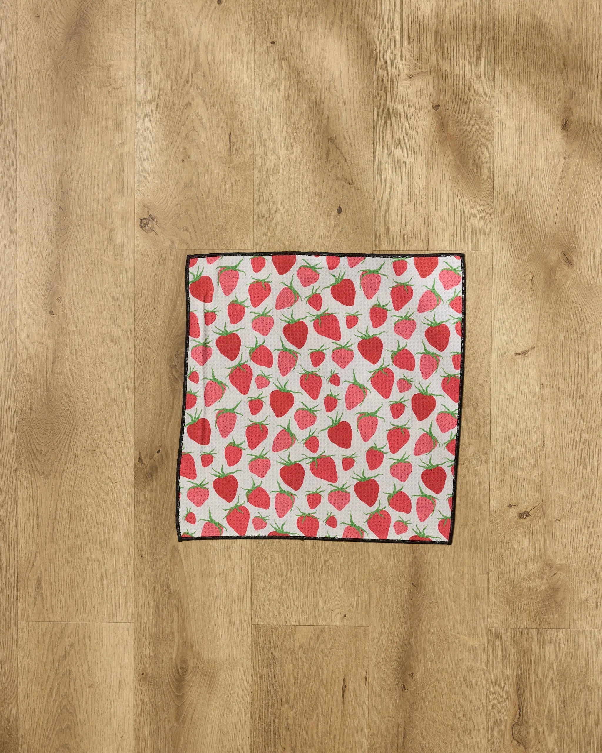 The Geometry "Strawberry Sniff" towel, a square ultra-absorbent pet towel with red strawberries and green leaves on white, is displayed flat on a light wood floor.