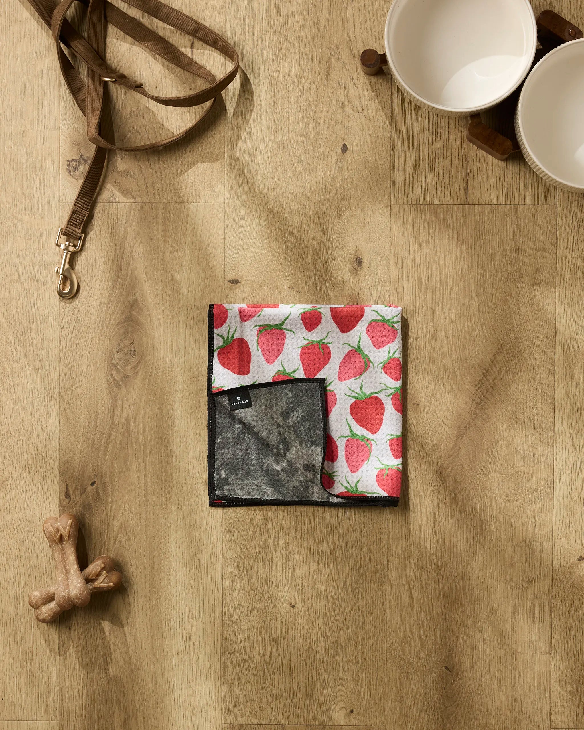 The Geometry Strawberry Sniff pet bandana is folded beside a brown leash, two white bowls on a wooden stand, a dog bone-shaped chew toy, and an ultra-absorbent pet towel for fast drying after playtime.