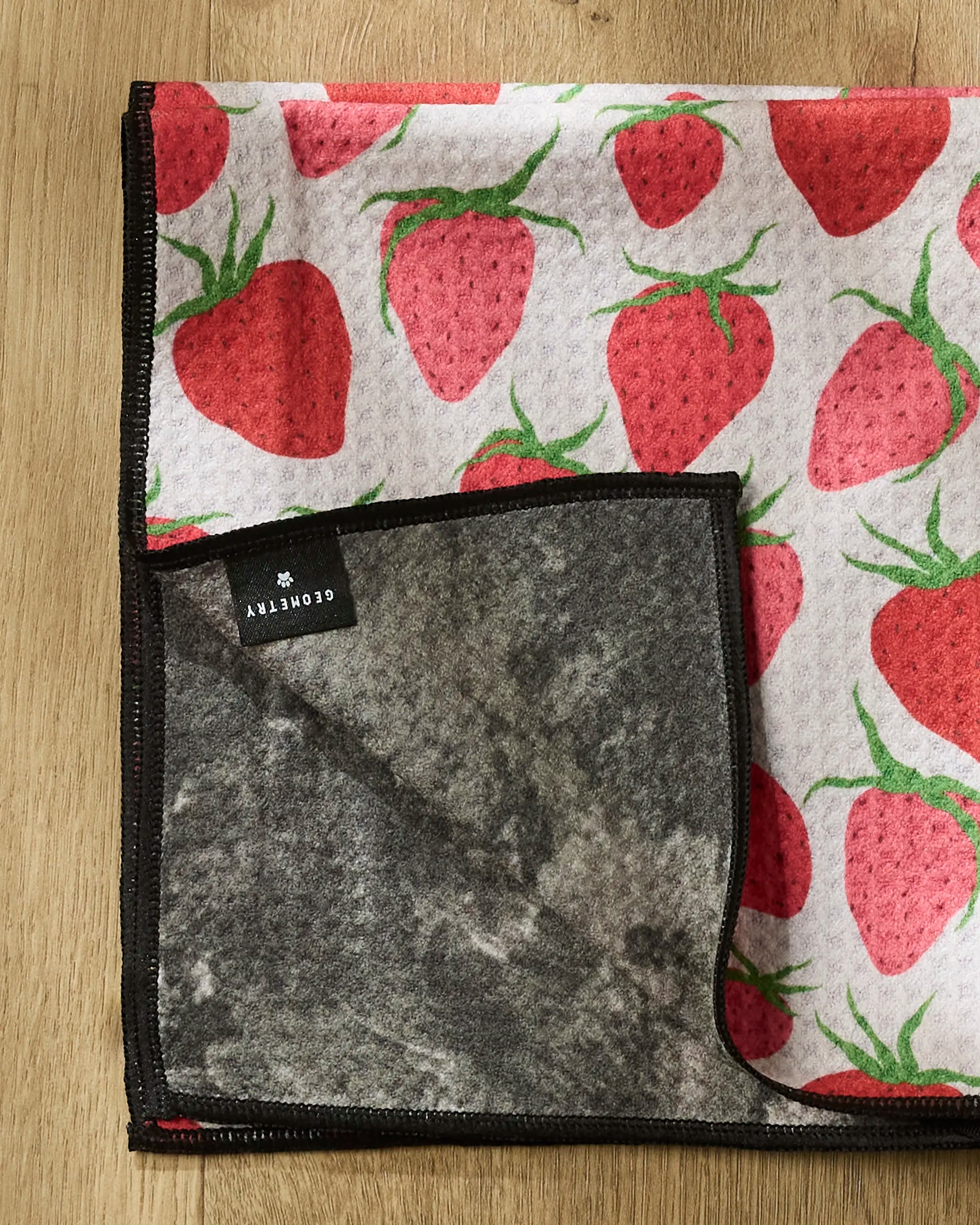 The Geometry Strawberry Sniff paw towel features a red strawberry pattern with green leaves. Displayed folded on wood, one corner is flipped to show its gray underside and a black label with white text.