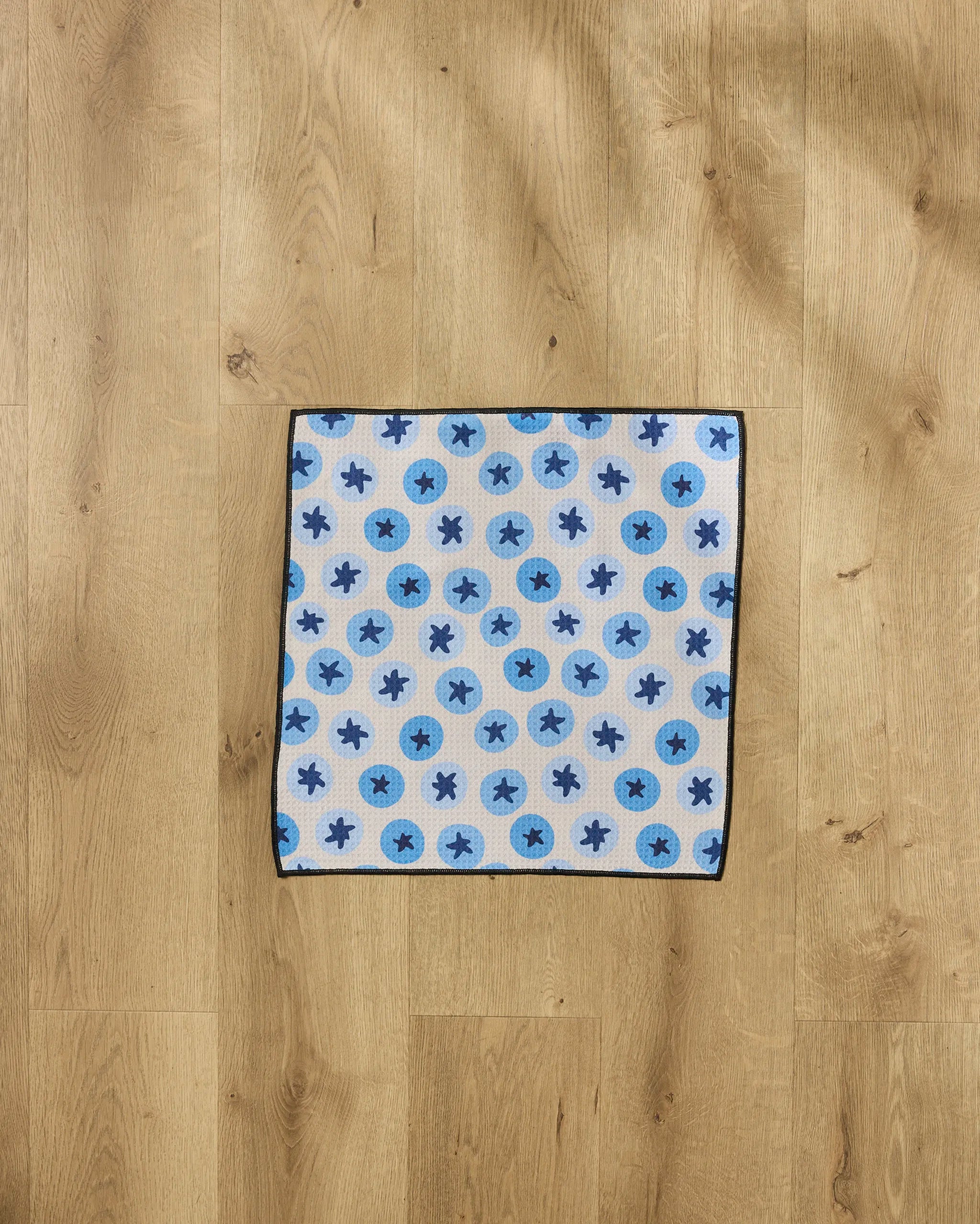 The Geometry Sweet Blueberries pet towel, a square ultra-absorbent towel with blue and dark blue circle and asterisk patterns, is laid flat on a light wooden floor.