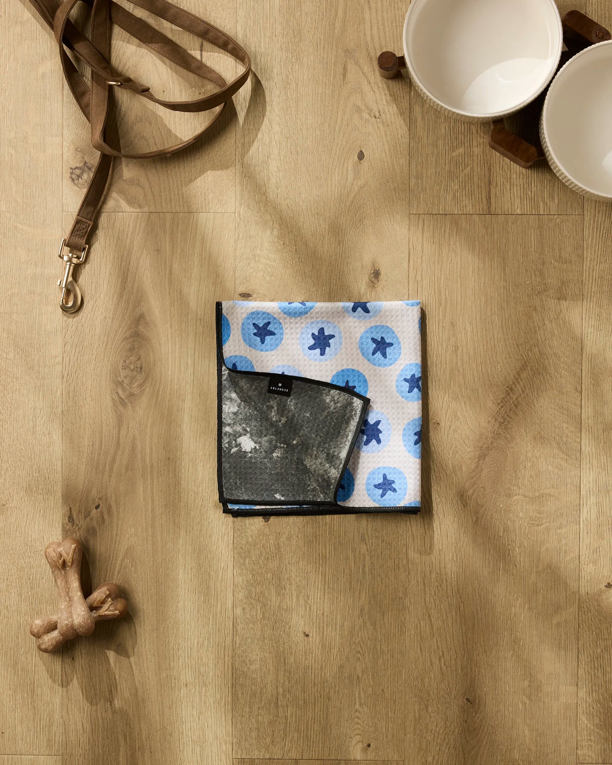 The Sweet Blueberries pet towel by Geometry, featuring blue paw prints, is folded alongside a brown leash, two white pet bowls on a wooden stand, and a dog bone toy on a light wood floor.