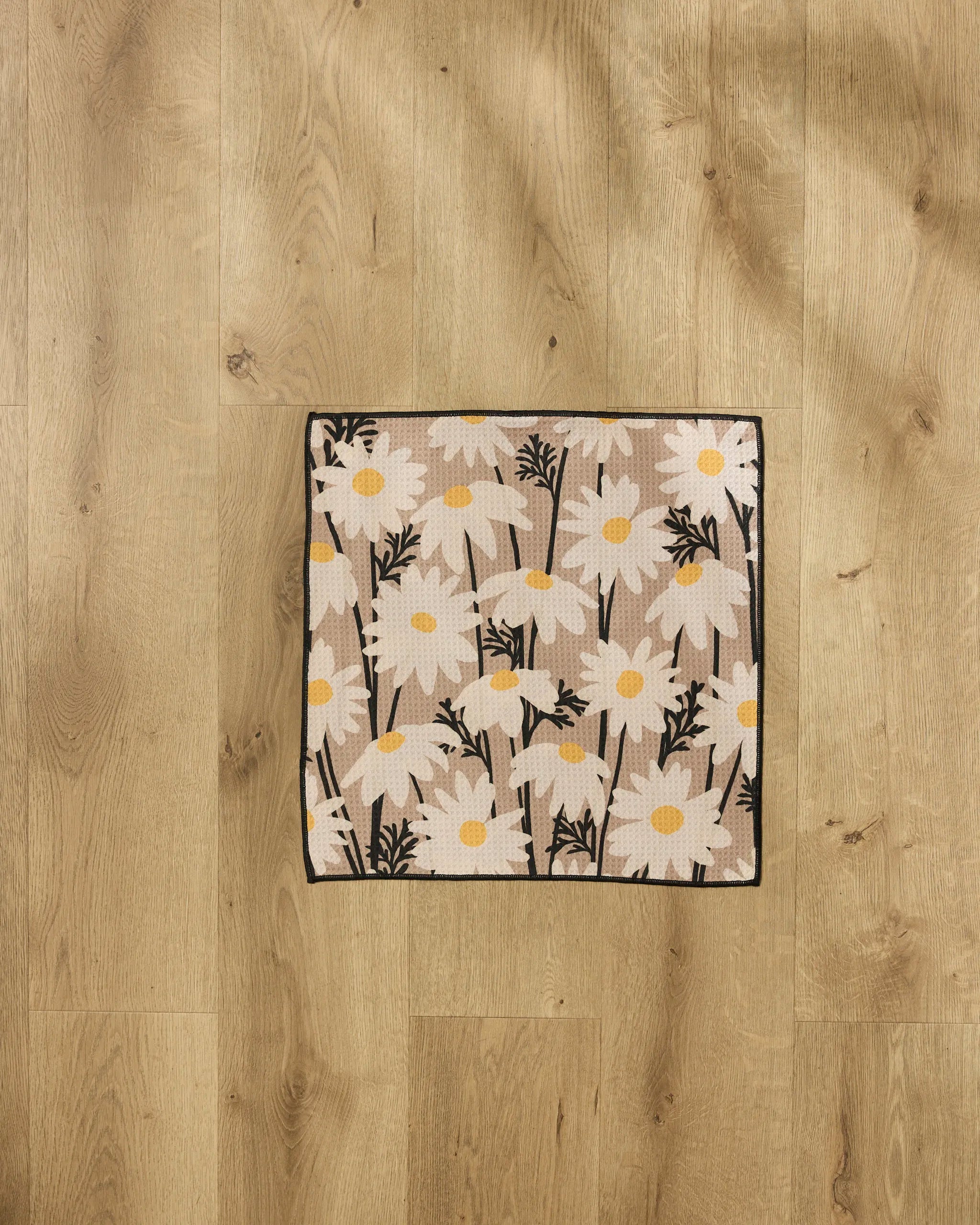 The Geometry Sweet Daisies recycled PET towel mat, featuring a square beige design with white daisies and yellow centers, bordered in black, is displayed on a light wood floor.