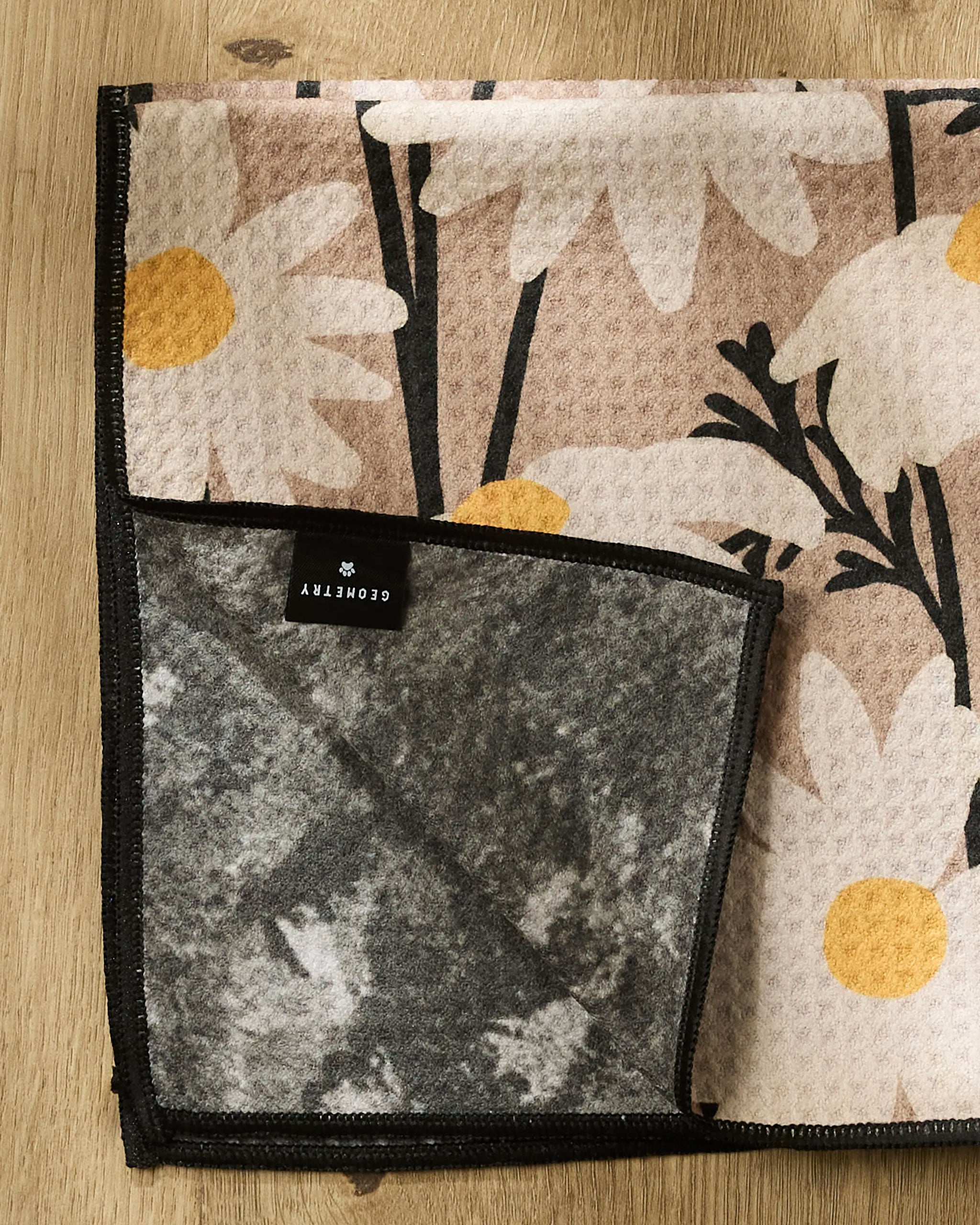 A beige and yellow Sweet Daisies towel by Geometry with a white daisy pattern lies on a wooden surface, one corner folded to show its gray underside. A black label with white "Geometry" text is visible.