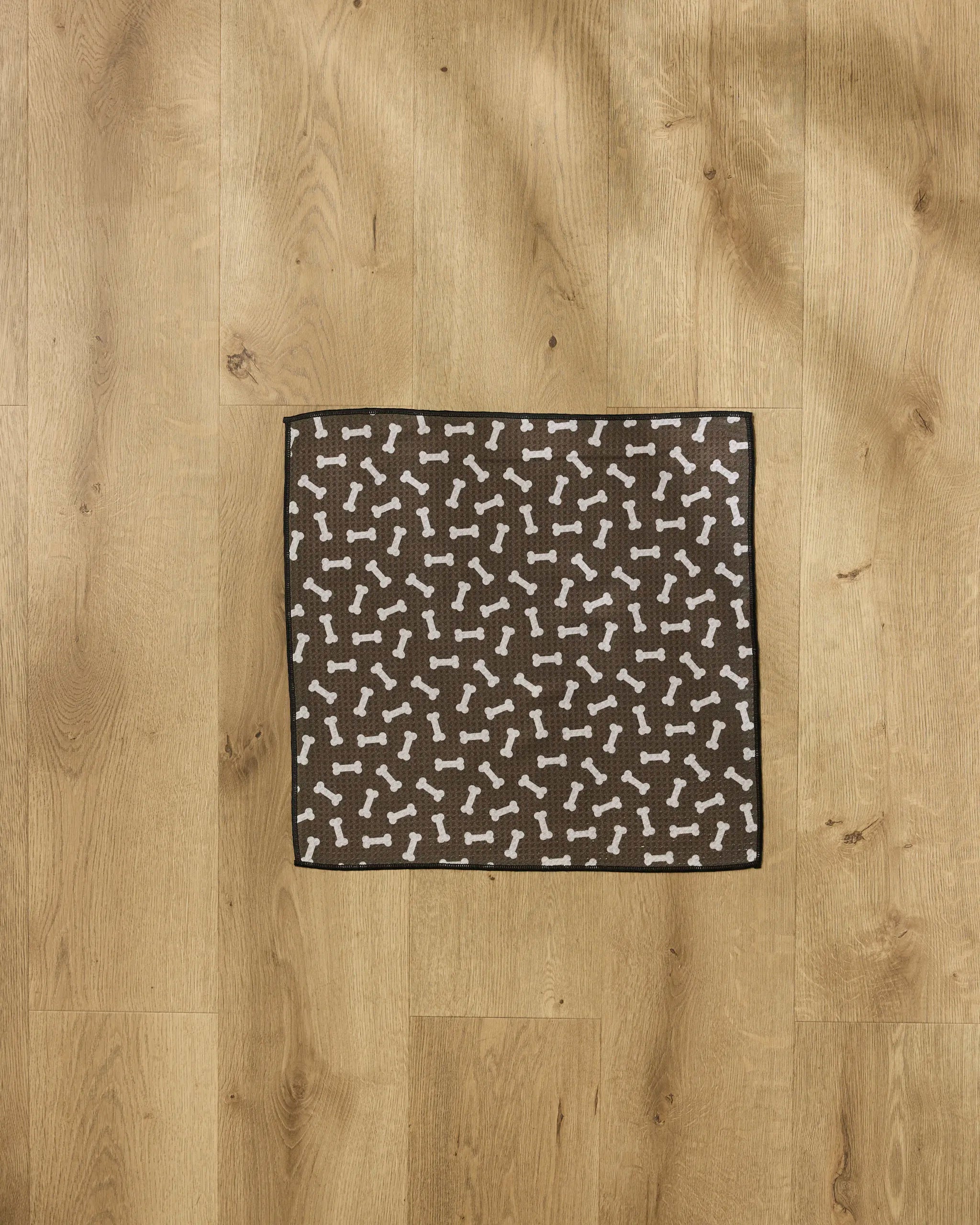 The Geometry Sweet Treats mat, featuring a brown square design with white dog bone patterns, sits on a light wood floor and doubles as an ultra-absorbent dog towel to keep your pet’s paws dry and clean.