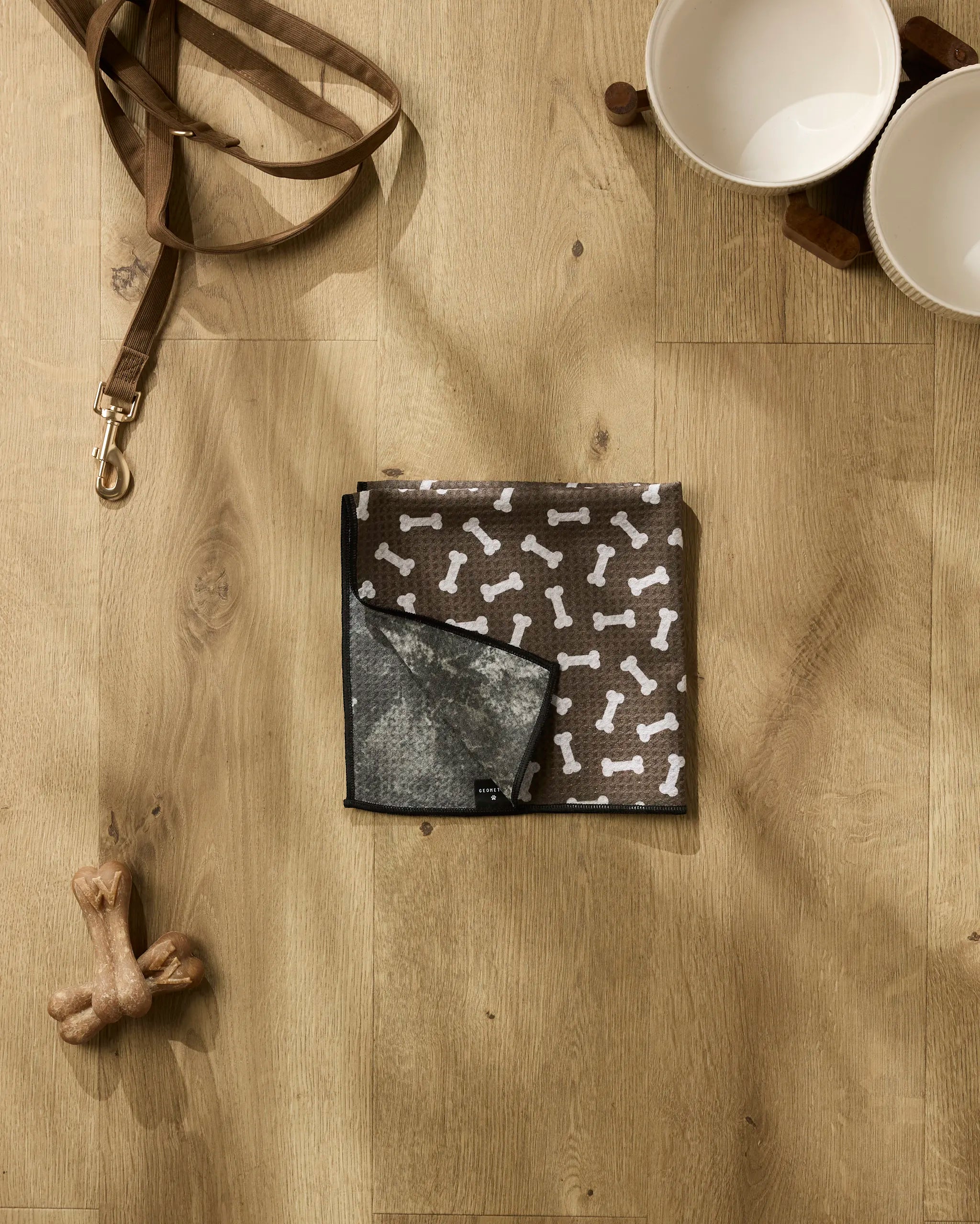 Geometry's Sweet Treats collection, including a bone-shaped toy, ultra-absorbent dog towel with white bone patterns, leash, and dual white bowls on a wooden stand, is arranged on a light wood floor.