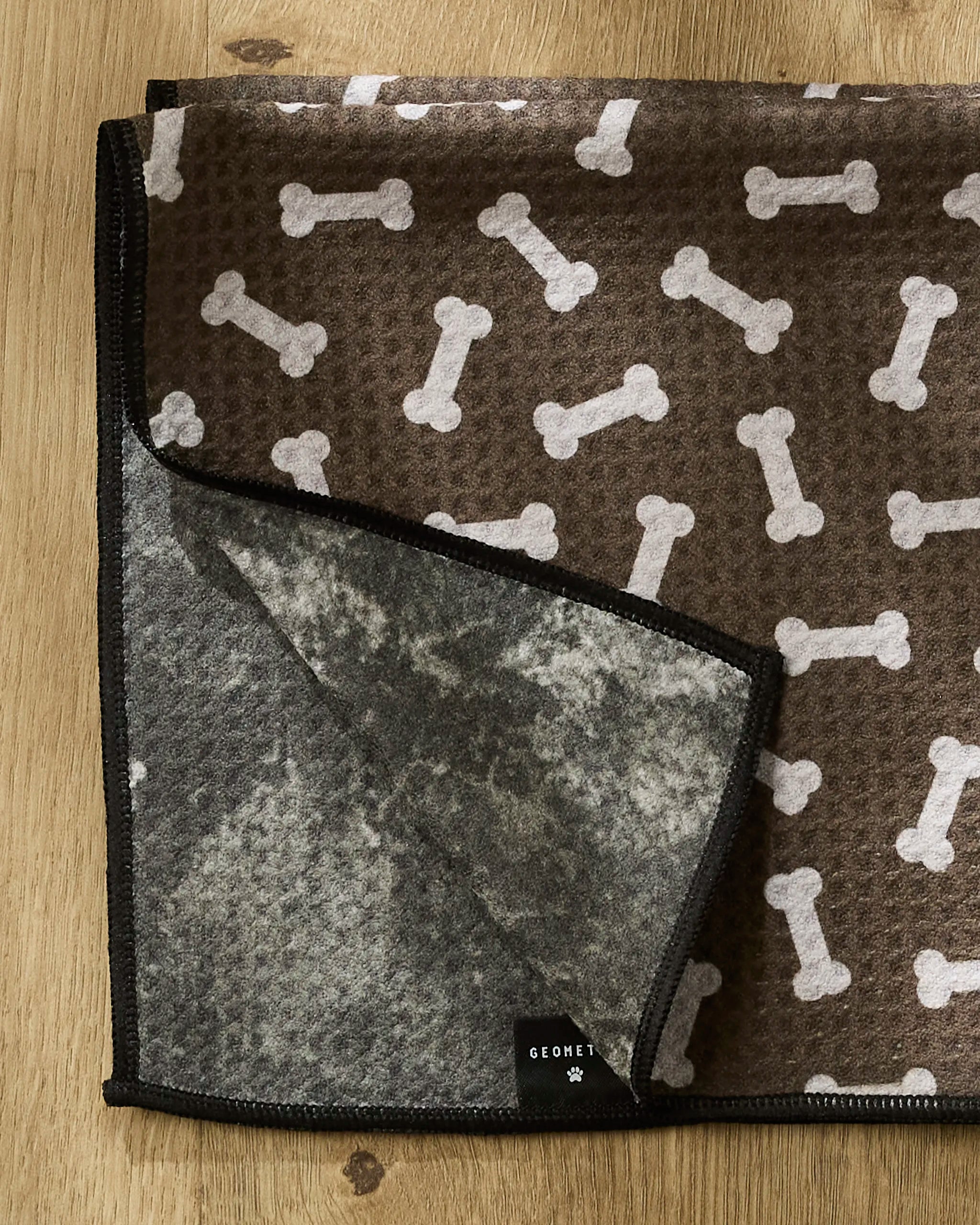 The Geometry Sweet Treats pet blanket, featuring a white bone pattern on brown and a gray underside with a black GEOMET label, rests on wood—ideal as an ultra-absorbent or recycled dog towel.