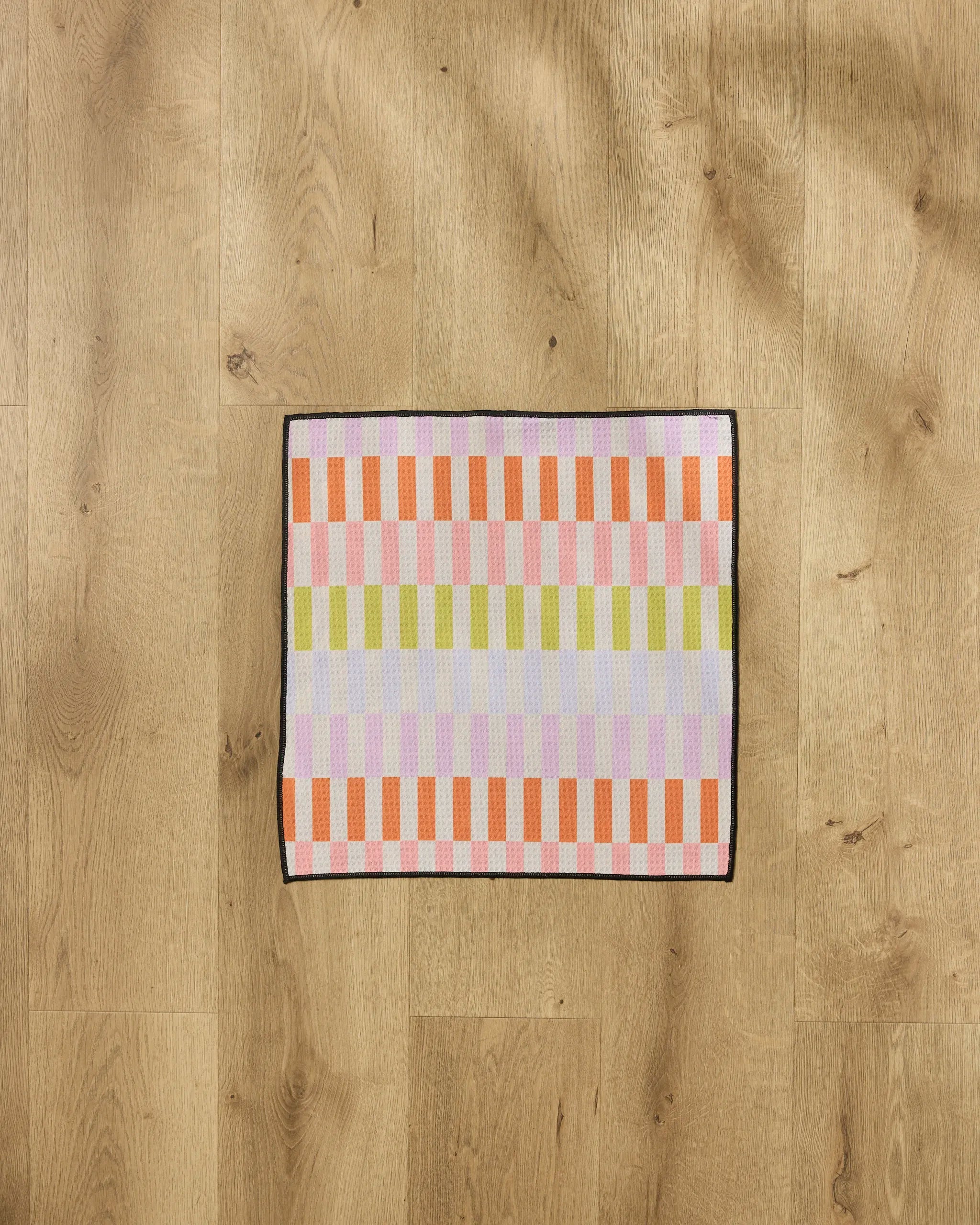 A small rectangular Wag Wise towel from Geometry, featuring a colorful orange, pink, purple, and green checkered pattern, sits on a light wooden floor.
