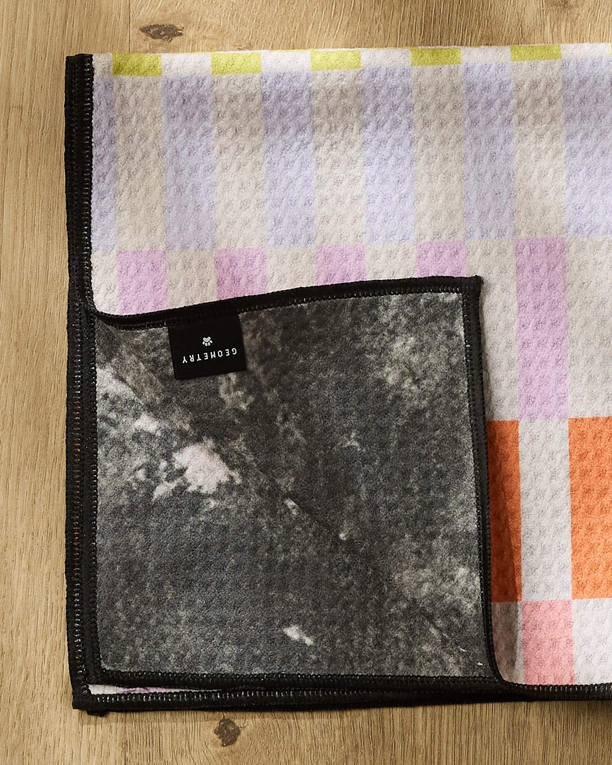 The Geometry Wag Wise Paw Towel features a soft, pastel checkered pattern and is folded on a wooden surface. A flipped corner reveals its dark marbled underside and a black label with white text.