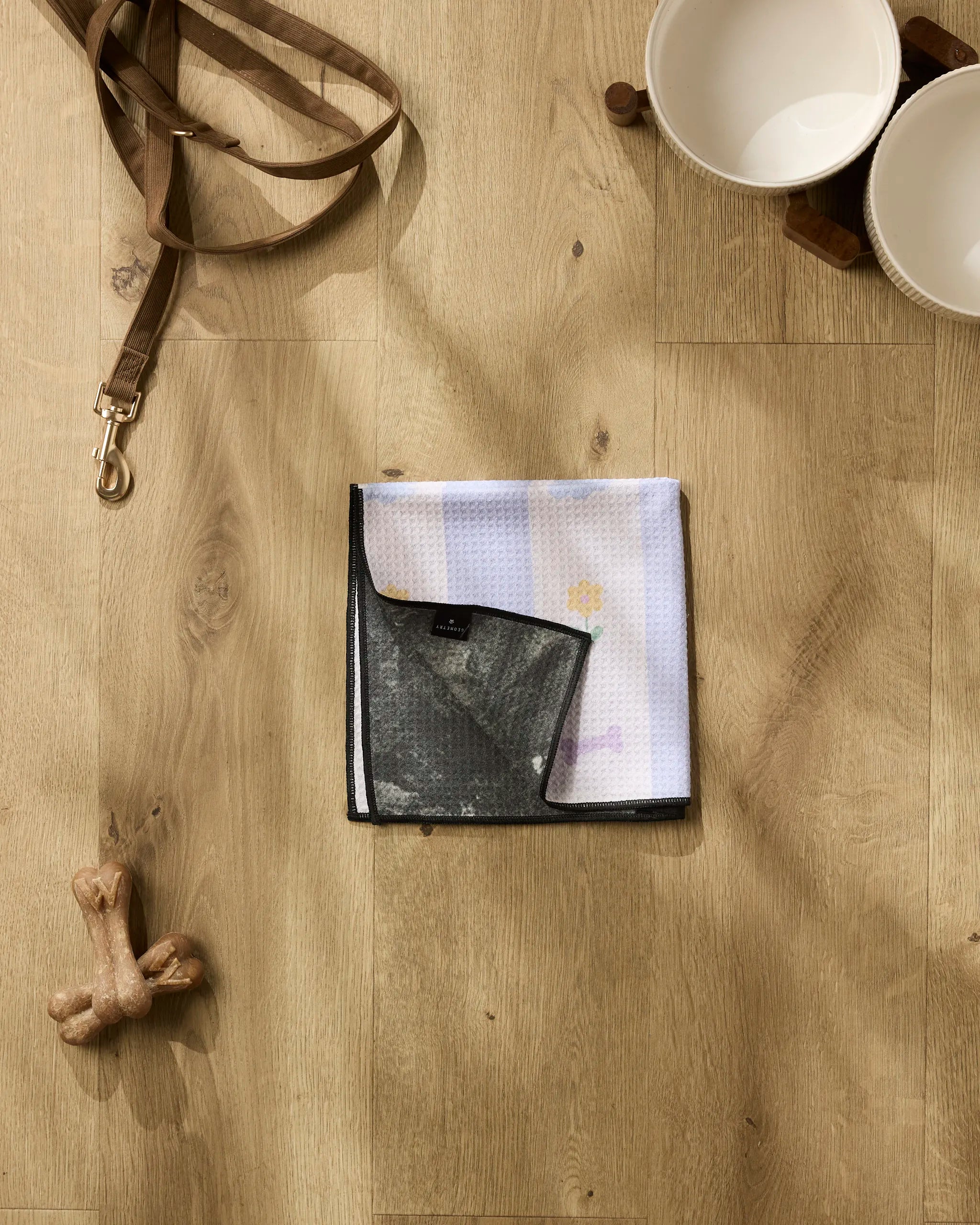 On a light wood floor, Wagging Blooms by Letters By Shells features a dog leash, two white pet bowls, a dog bone toy, and an ultra-absorbent Paw Towel with a black border.
