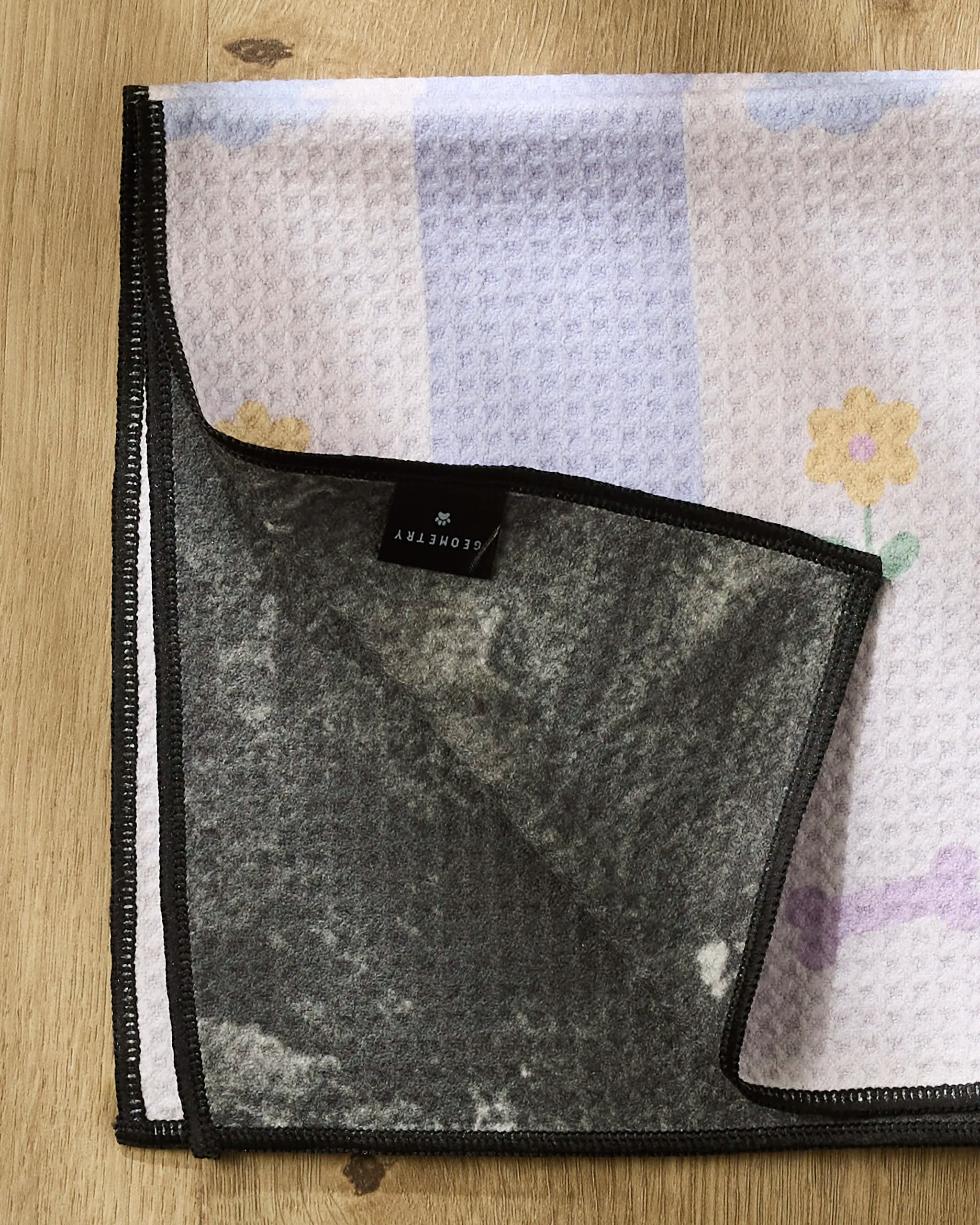 The Wagging Blooms Paw Towel by Letters By Shells, featuring pastel floral patterns, is displayed on a wooden surface with a corner flipped to show its dark ultra-absorbent lining and attached care label.