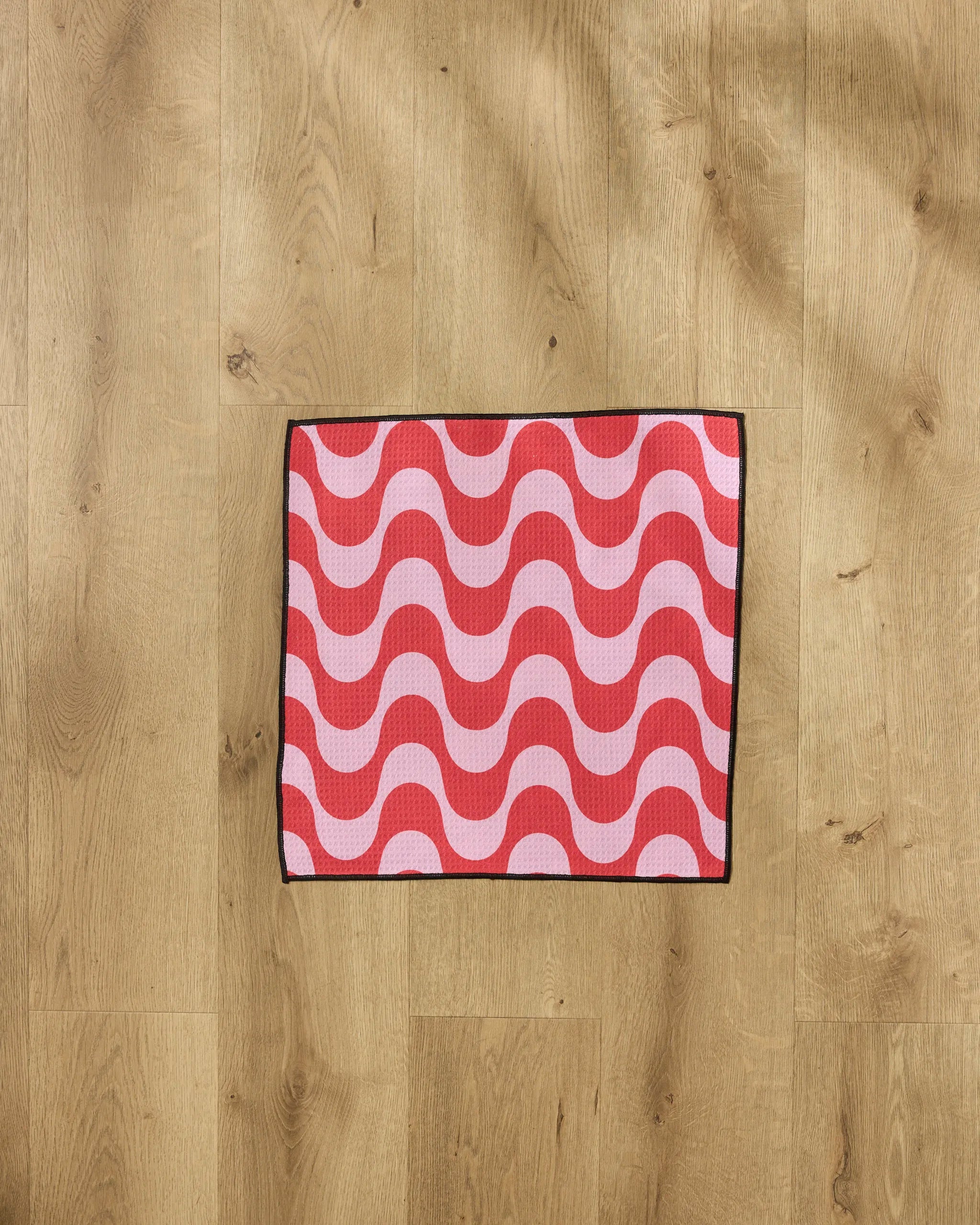 The Geometry Waves mat, featuring a red and pink wavy pattern with a black border, rests on a light wood floor—an ultra-absorbent and stylish paw towel for your pet.