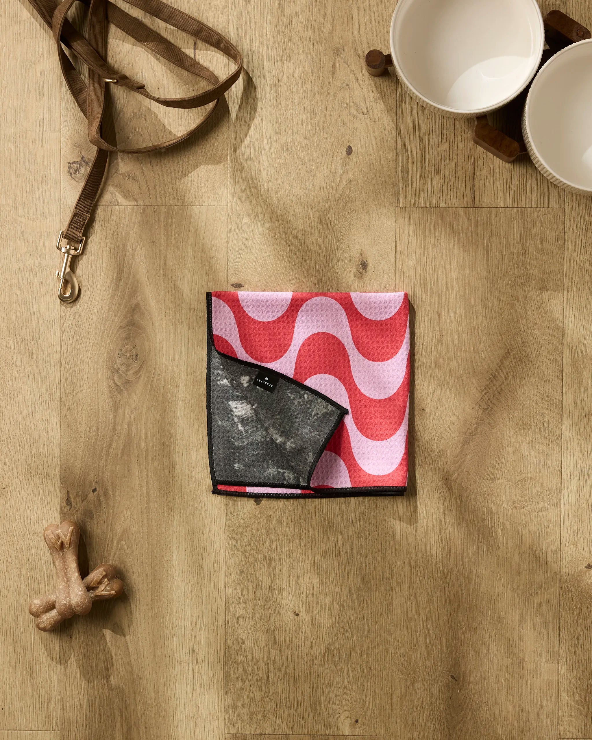 The Geometry Waves bandana, featuring a pink and white wavy pattern, is folded on a wooden floor beside a leather leash, white dog bowls, a dog bone toy, and an ultra-absorbent paw towel for quick post-walk cleanups.