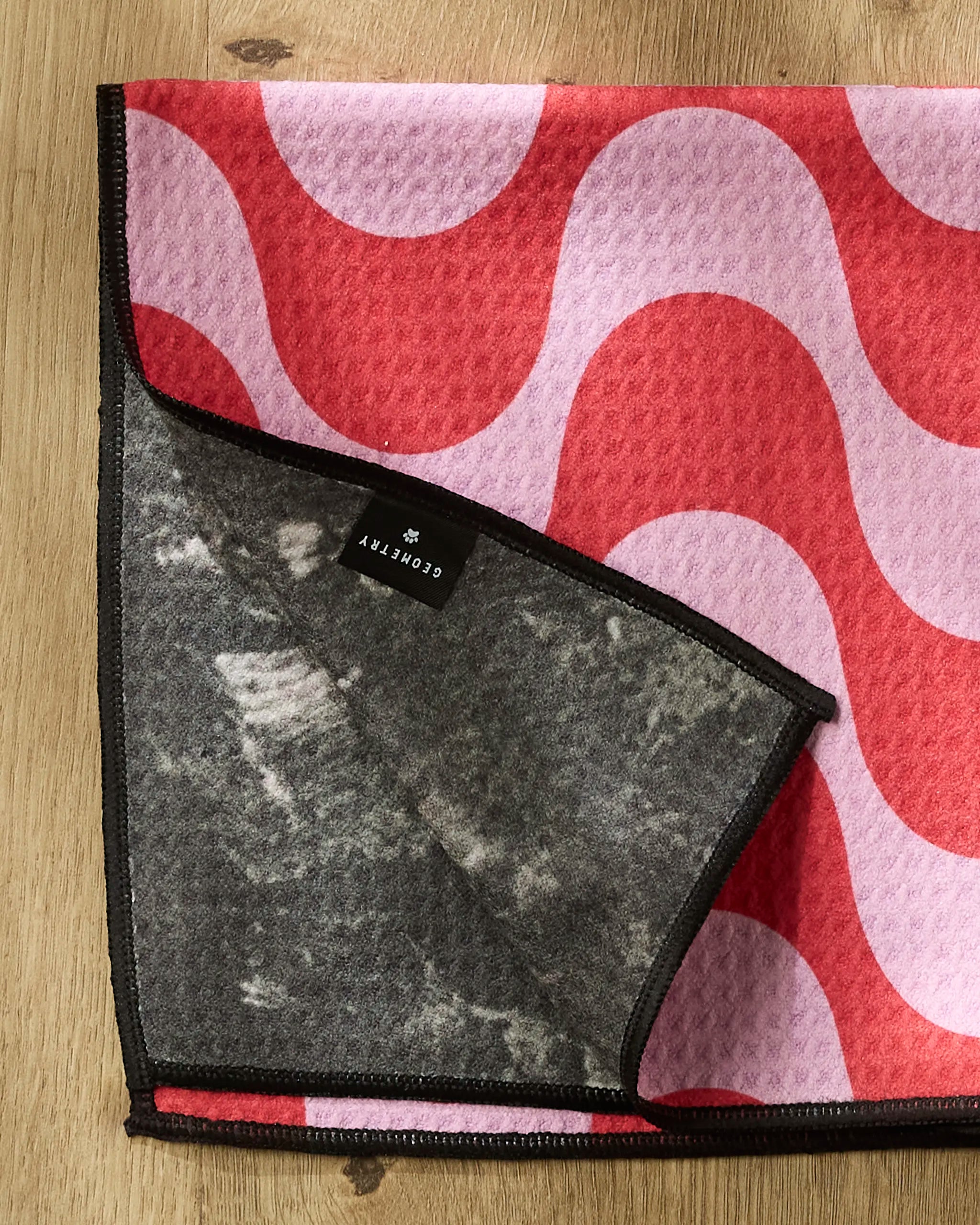 A textured, ultra-absorbent Geometry towel called "Waves," featuring a pink and red wavy pattern, lies on a wooden surface. A flipped corner reveals its gray underside and a black tag labeled CONCRETE AF.