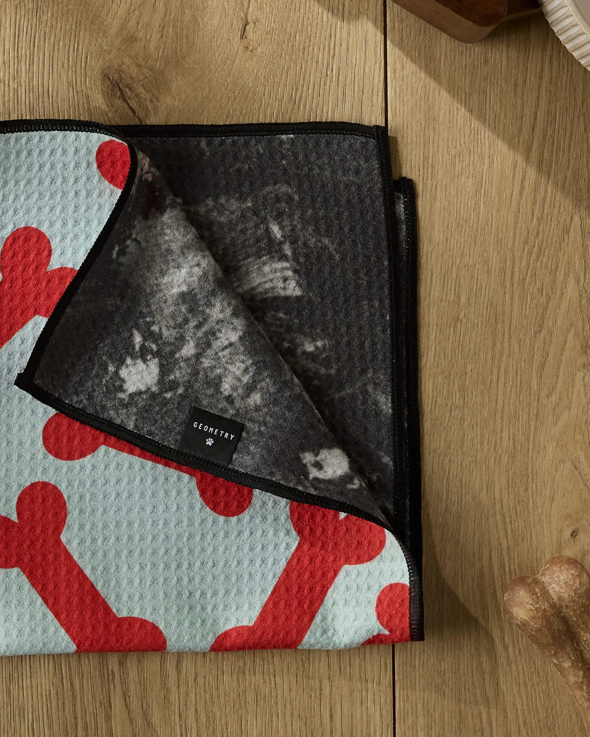 A folded Geometry Yuletide Pup dog towel with large red dog bones on a light background rests on wood, a corner flipped to show its dark, textured underside and a small Geometry brand tag.