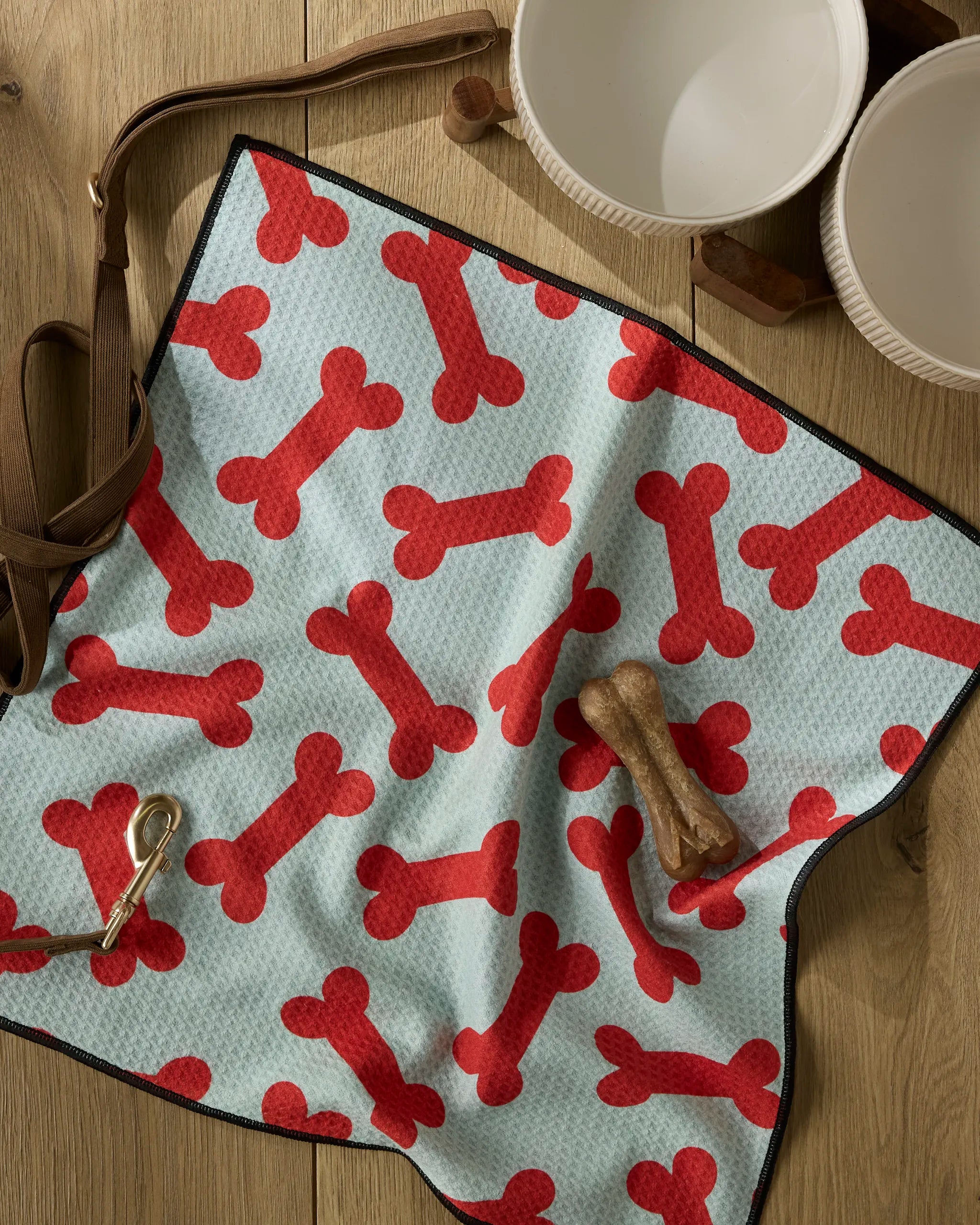 The Geometry Yuletide Pup mat in light blue with red dog bone pattern rests on a wooden floor, accessorized with a brown leash, dog bone treat, collar, ultra-absorbent dog towel, and two white bowls nearby.